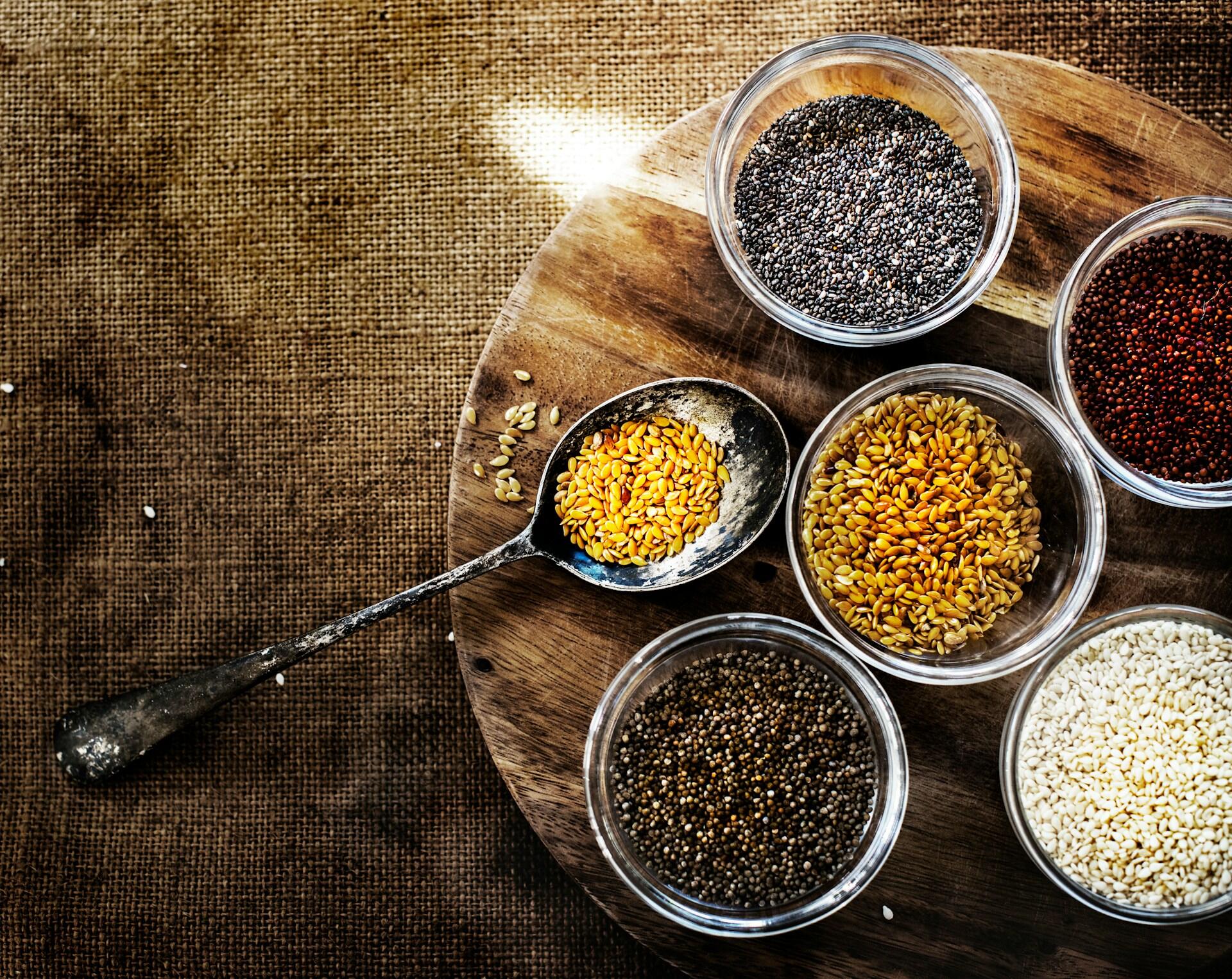 small bowls of different types of seeds on a wooden plate, a rustic spoon has a scoop of flax seeds in it