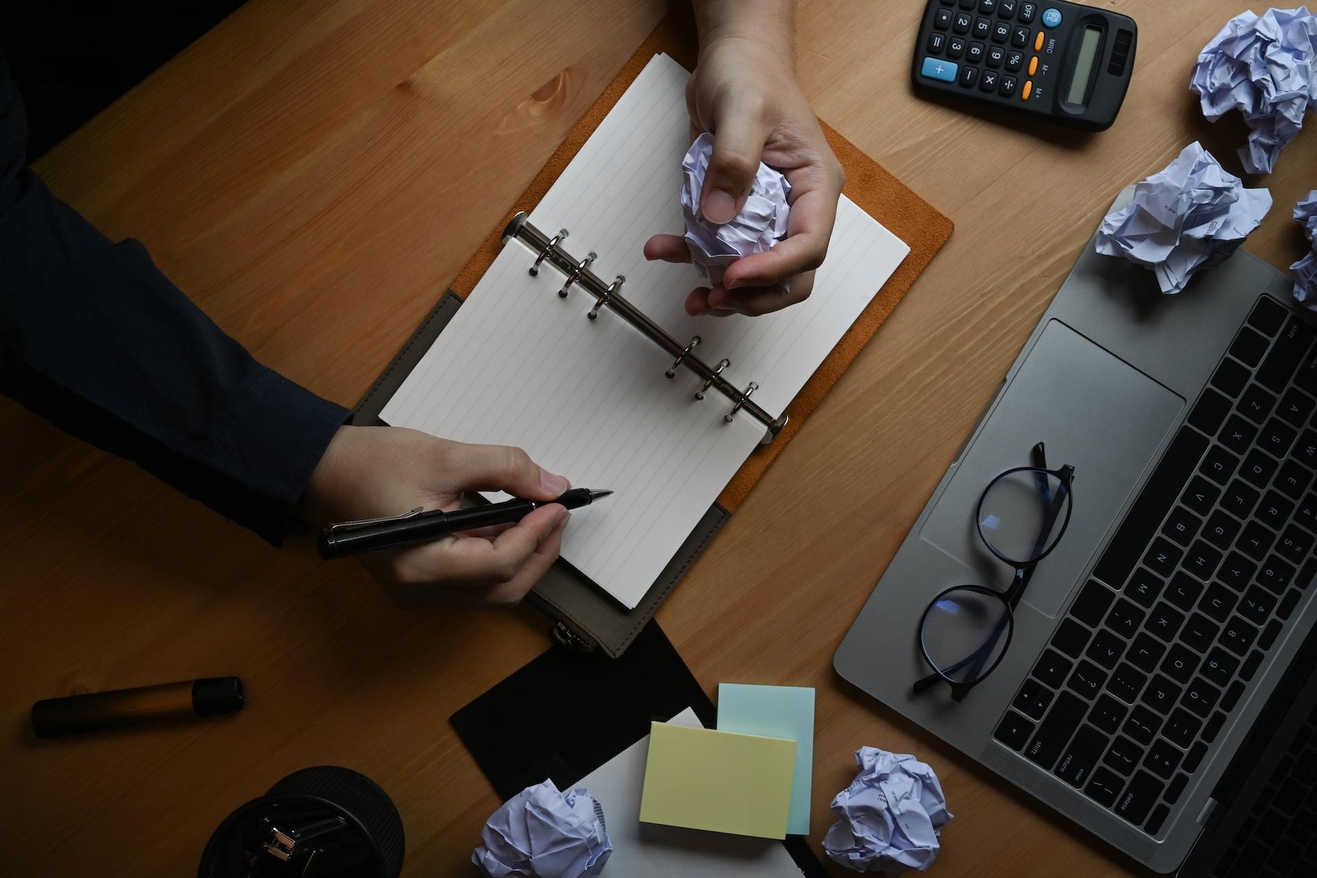 An overhead view of a person wearing a dark long sleeved top sitting at a wooden topped surface, writing in a spiral notepad in front of an open laptop computer, with several wads of discarded paper scattered around them.