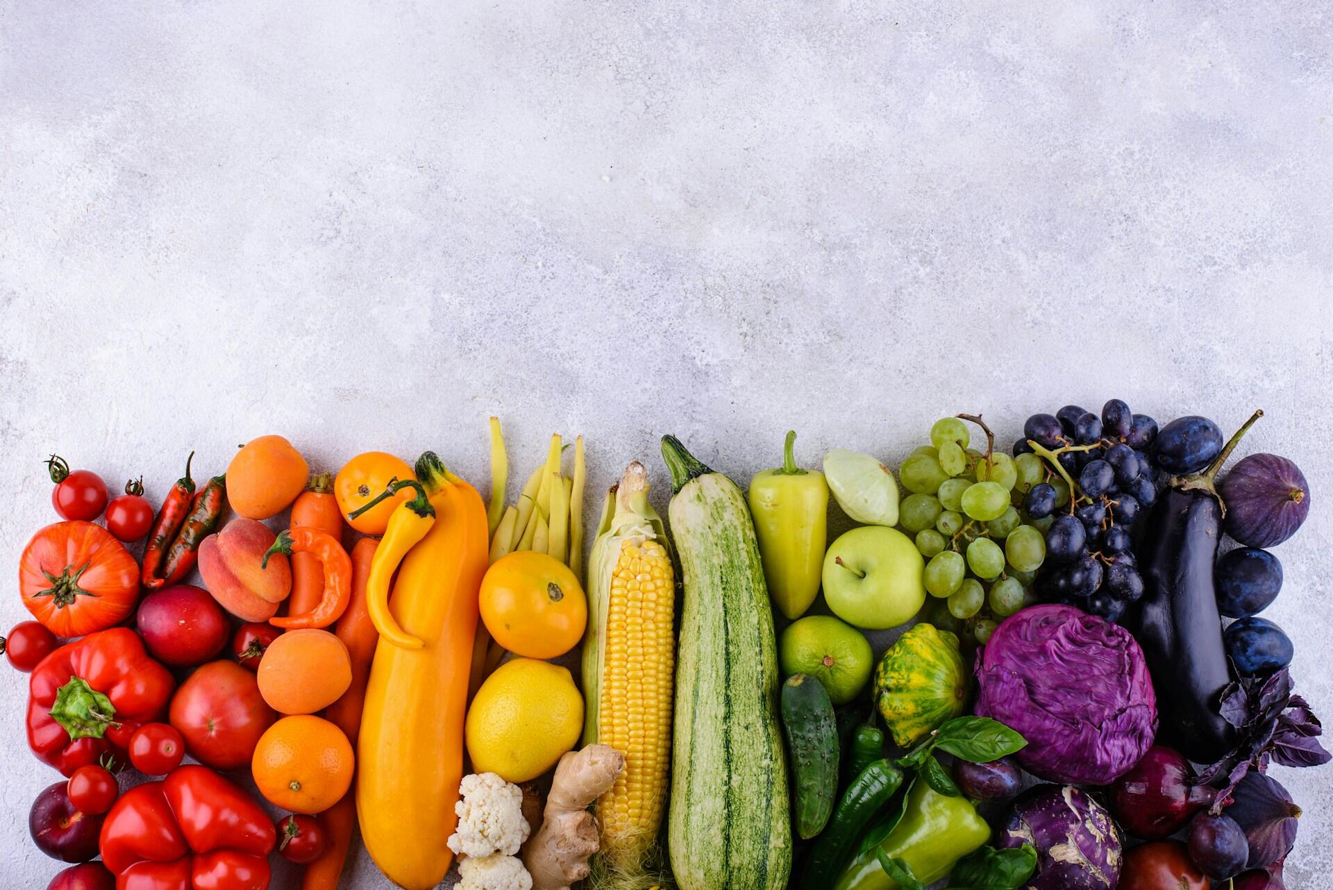 a rainbow of fruits and vegetables including tomatoes, oranges, lemons, corn, peppers, zucchini, apples, grapes, cabbage, and aubergine