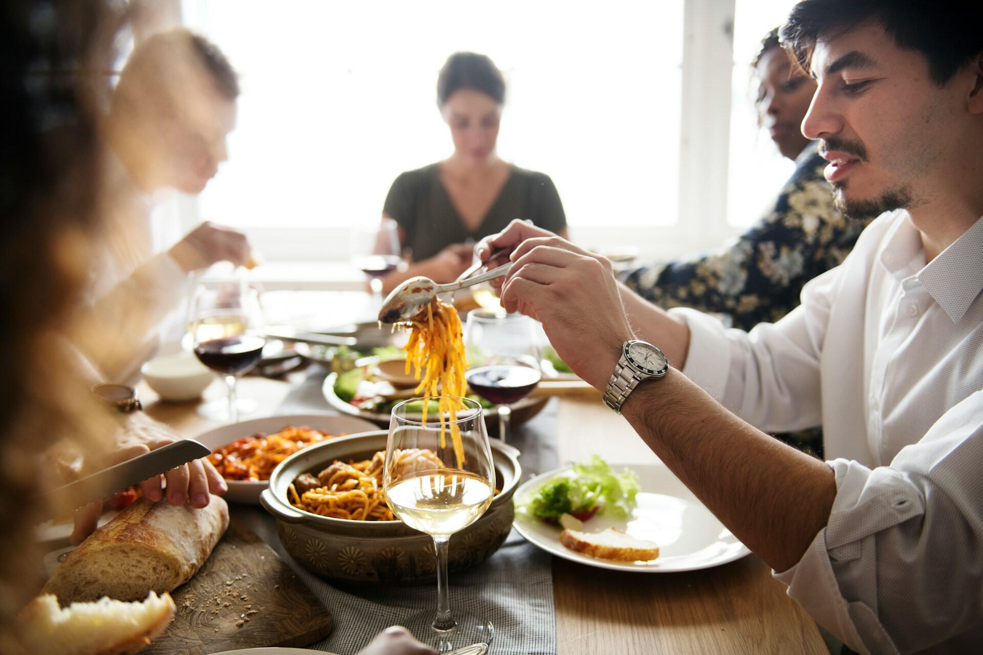 people at a dinner party, where one guest takes a serving of pasta with red sauce and puts it on their plate along with a small salad and slice of bread