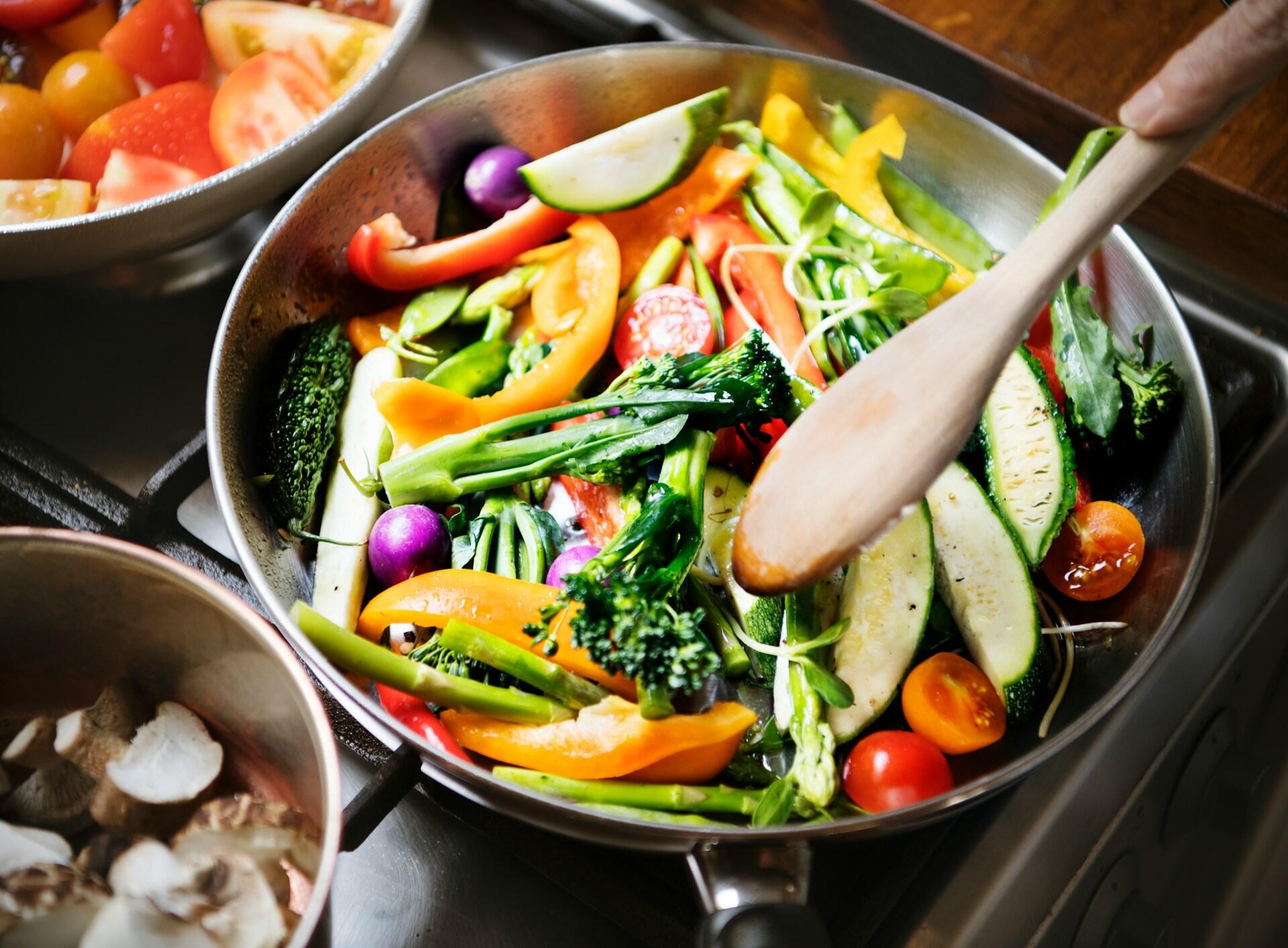 steel frying pan filled with a variety of veggies, cooking on the stove top being stirred by a wooden spoon. a pot of mushrooms and tomatoes cook on the adjacent burners