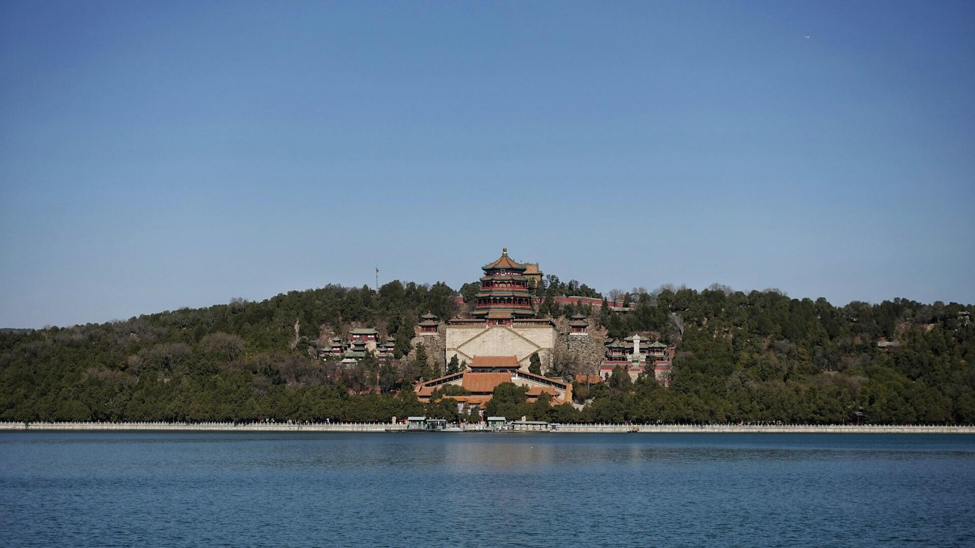Historic multi-tiered building atop a forested hill, overlooking a calm lake with clear blue sky.