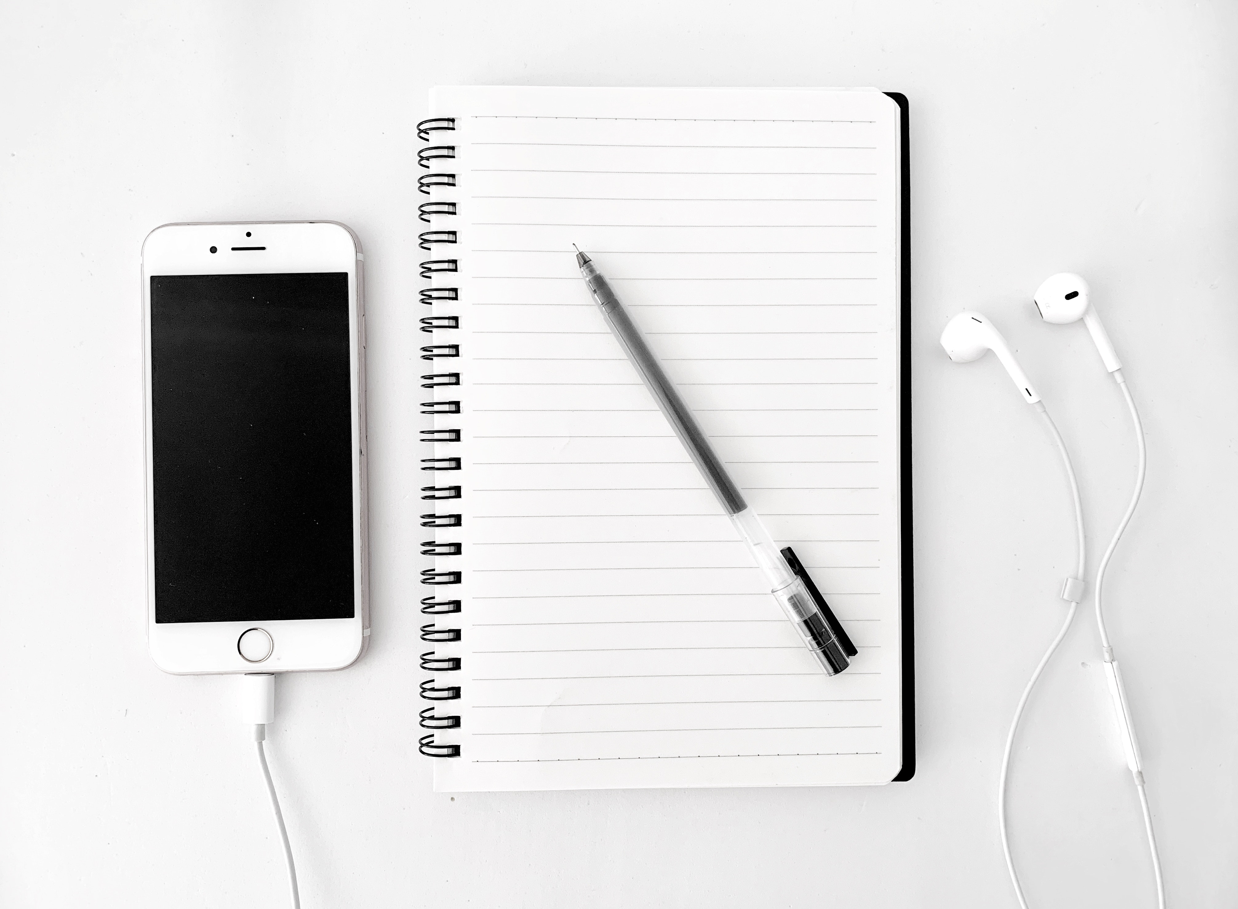 A white mobile phone, a spiral notepad opened to a blank page, a black pen laying on that page and a white set of earbuds, all set against a white background.
