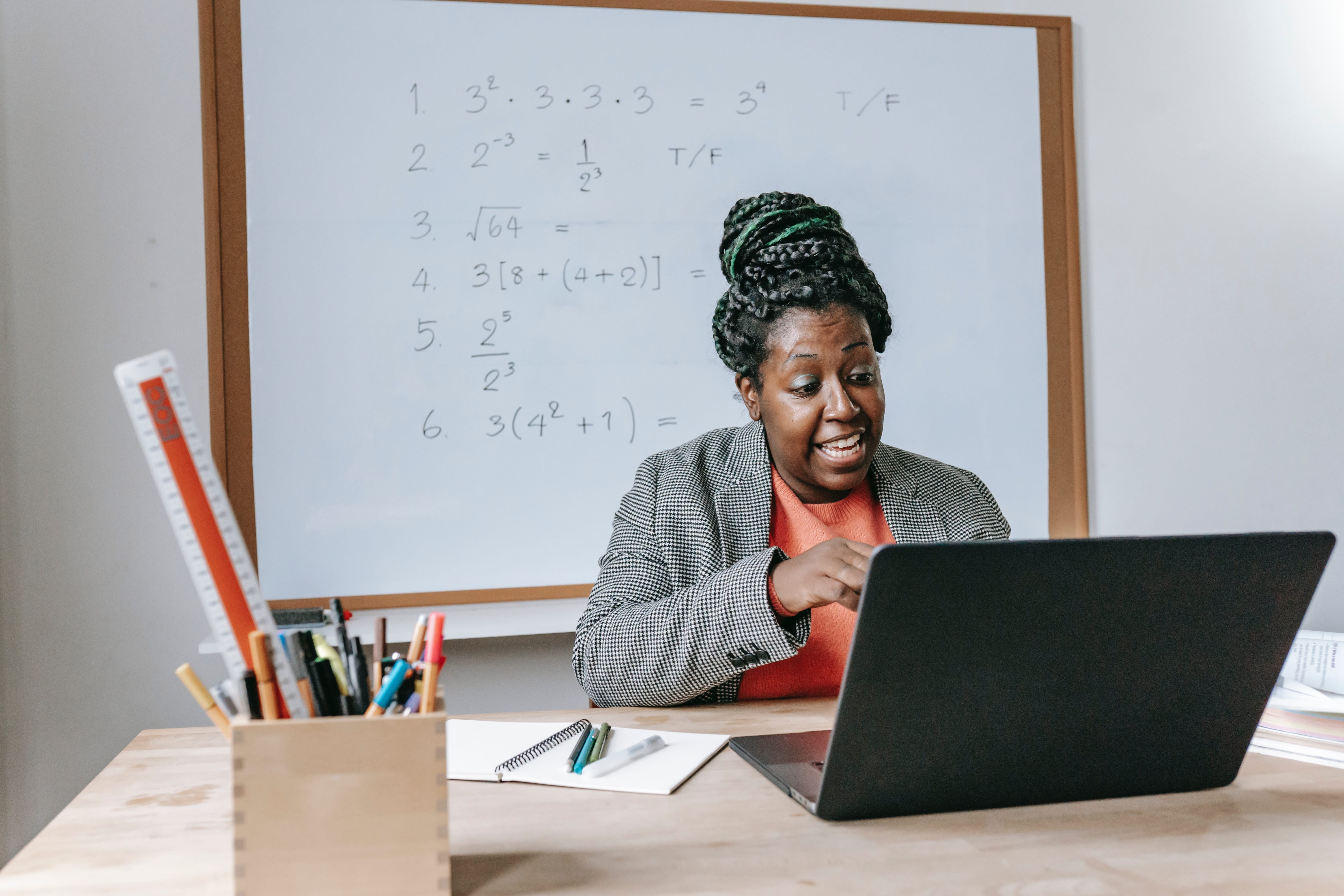 A person wearing an orange top under a grey blazer sits at a blond wood table bearing a wooden pencil cup full of writing implements and a long ruler, a notepad with several pens on it and an open laptop computer, at which the person looks, smiling.
