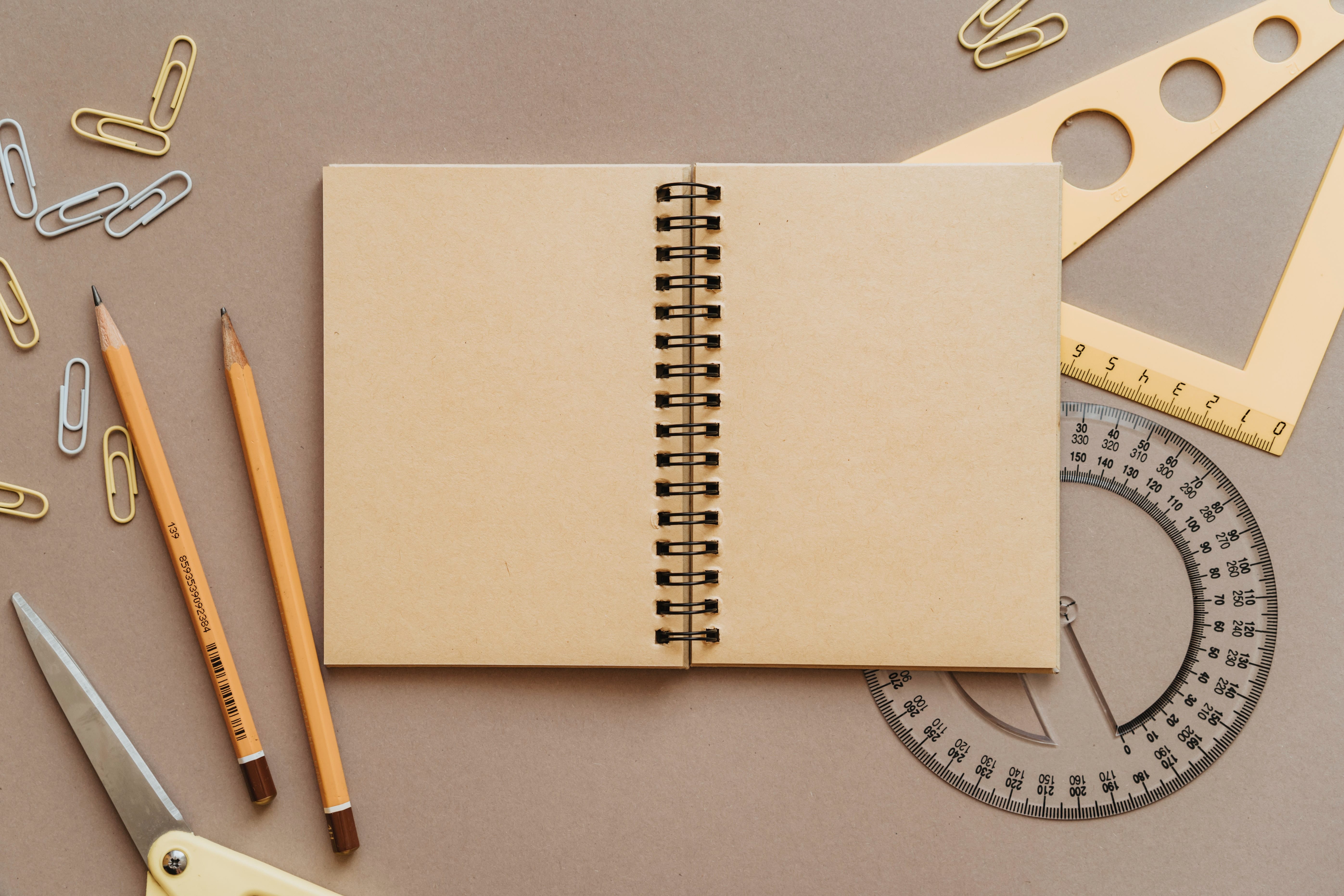 An open spiral-bound notebook displaying blank pages, surrounded by a protractor, a square, two pencils and some paperclips