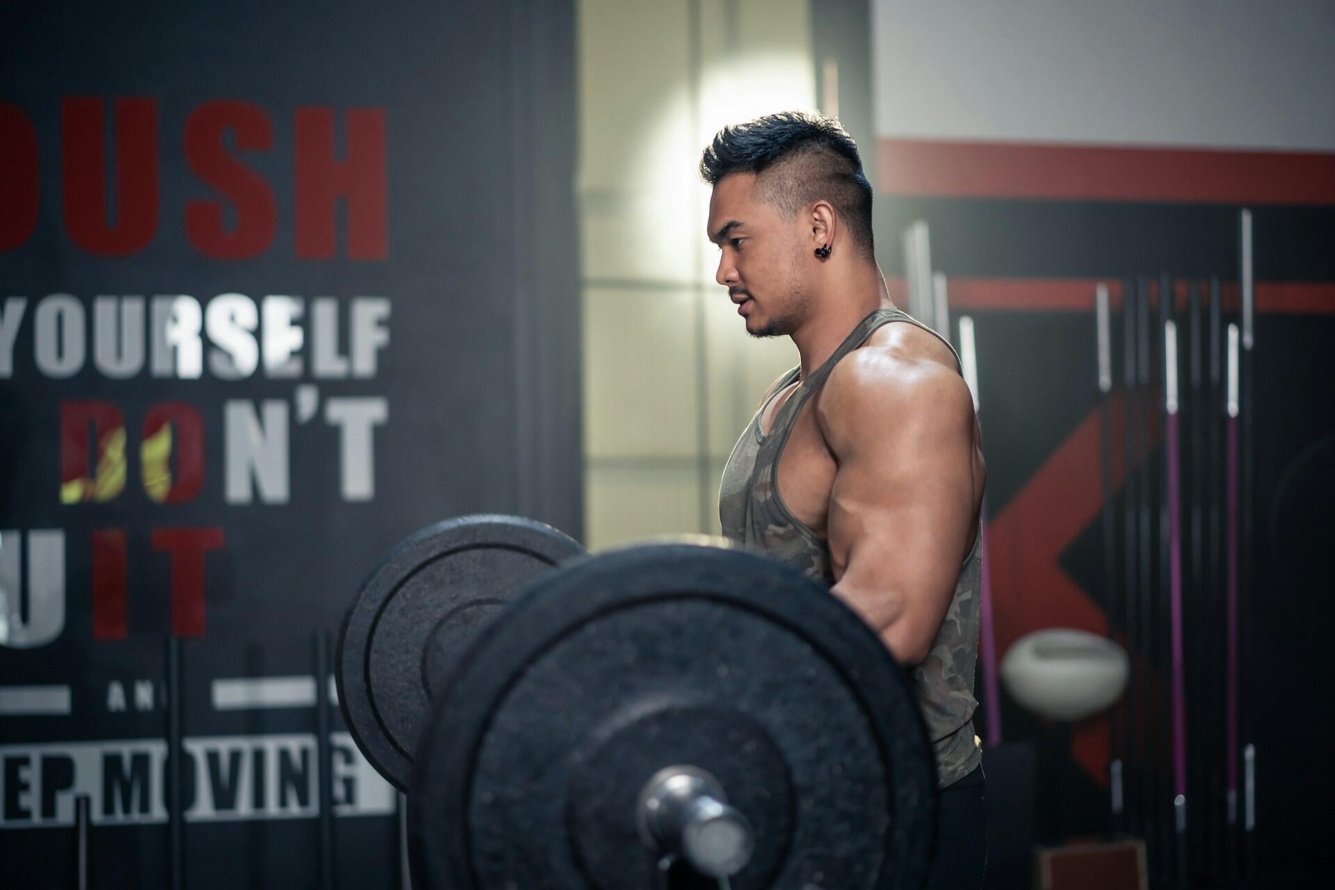 A muscular individual is lifting a heavy barbell in a gym, with motivational workout quotes displayed on the wall behind.