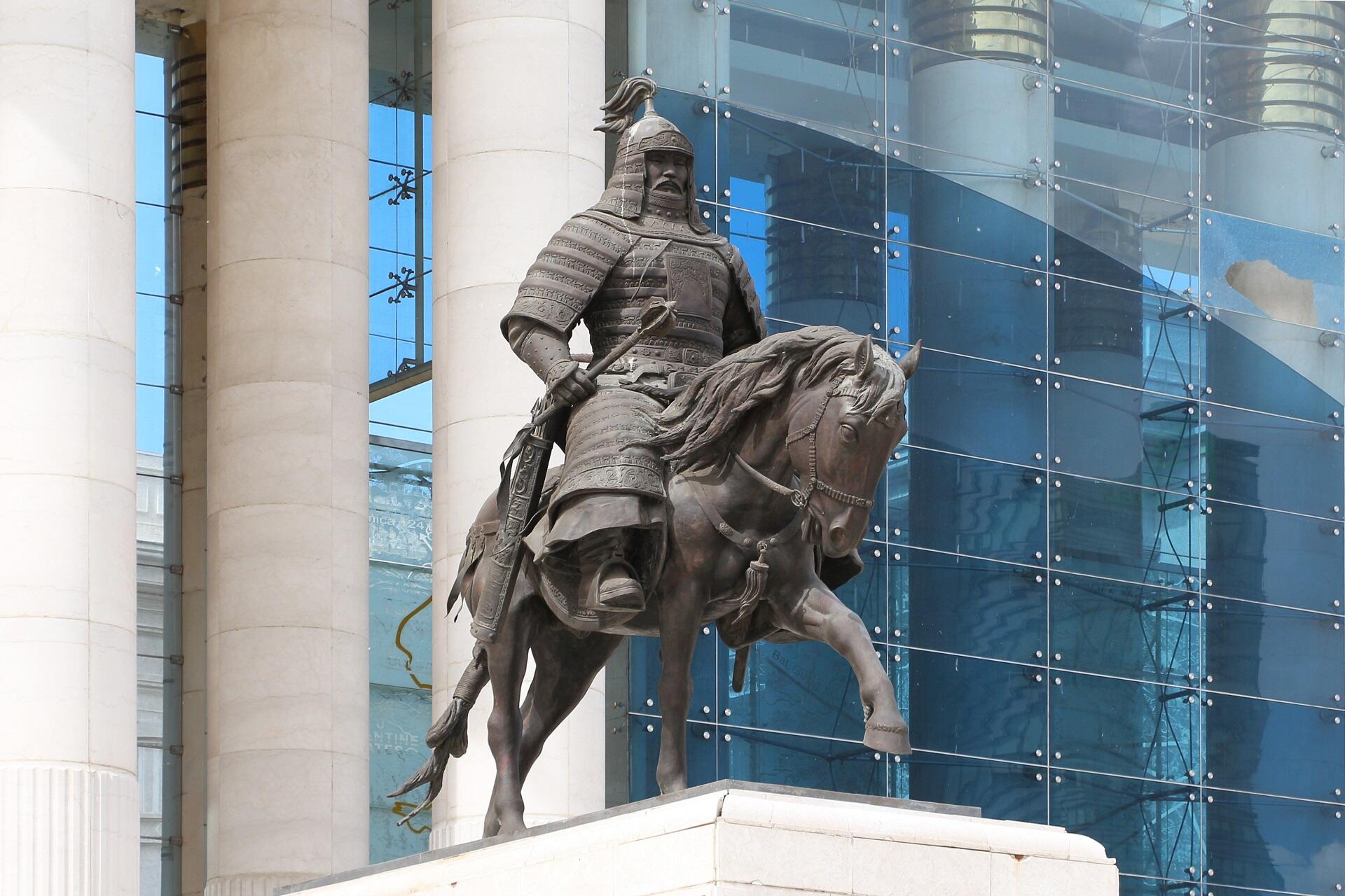 A statue of a man with a moustache wearing armour astride a horse in front of a glass-fronted building.