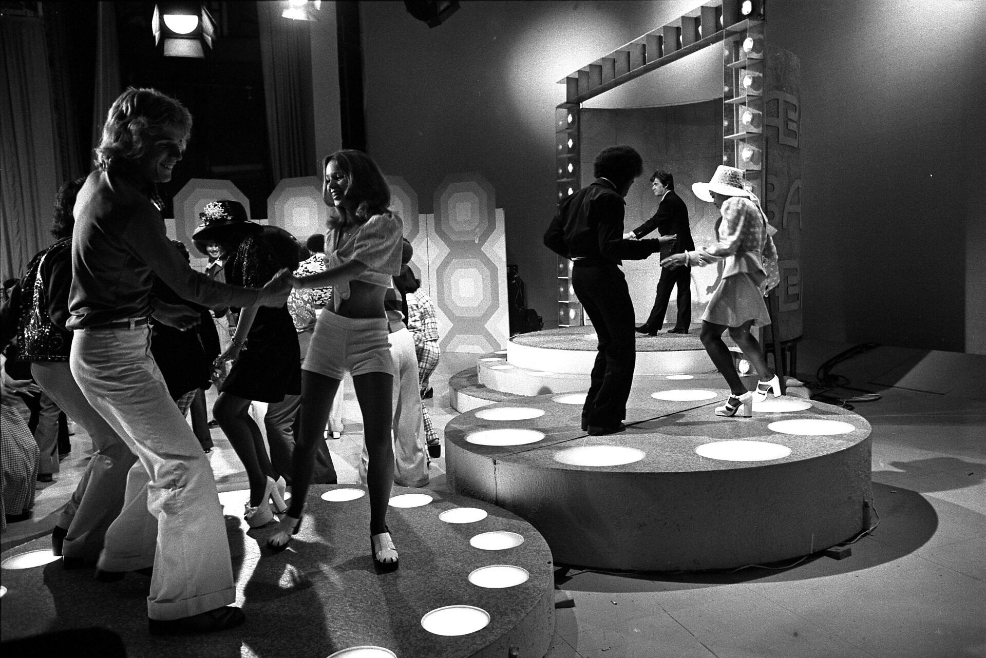 People in casual clothing dancing on a sound stage while a man in a suit looks on.