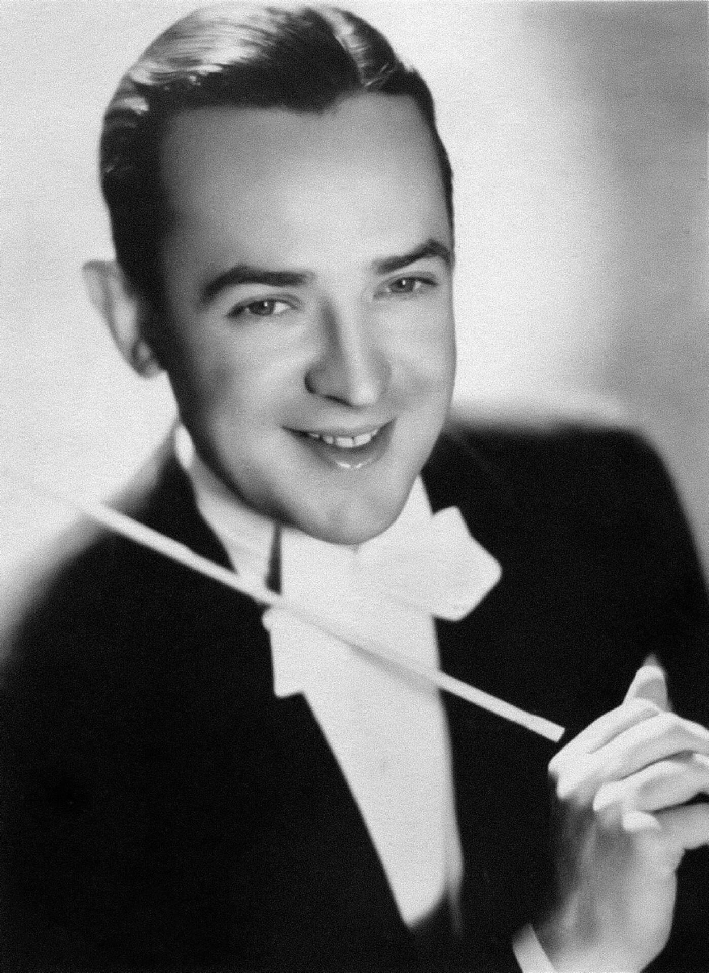 A smiling man wearing a formal suit and holding an orchestra baton.