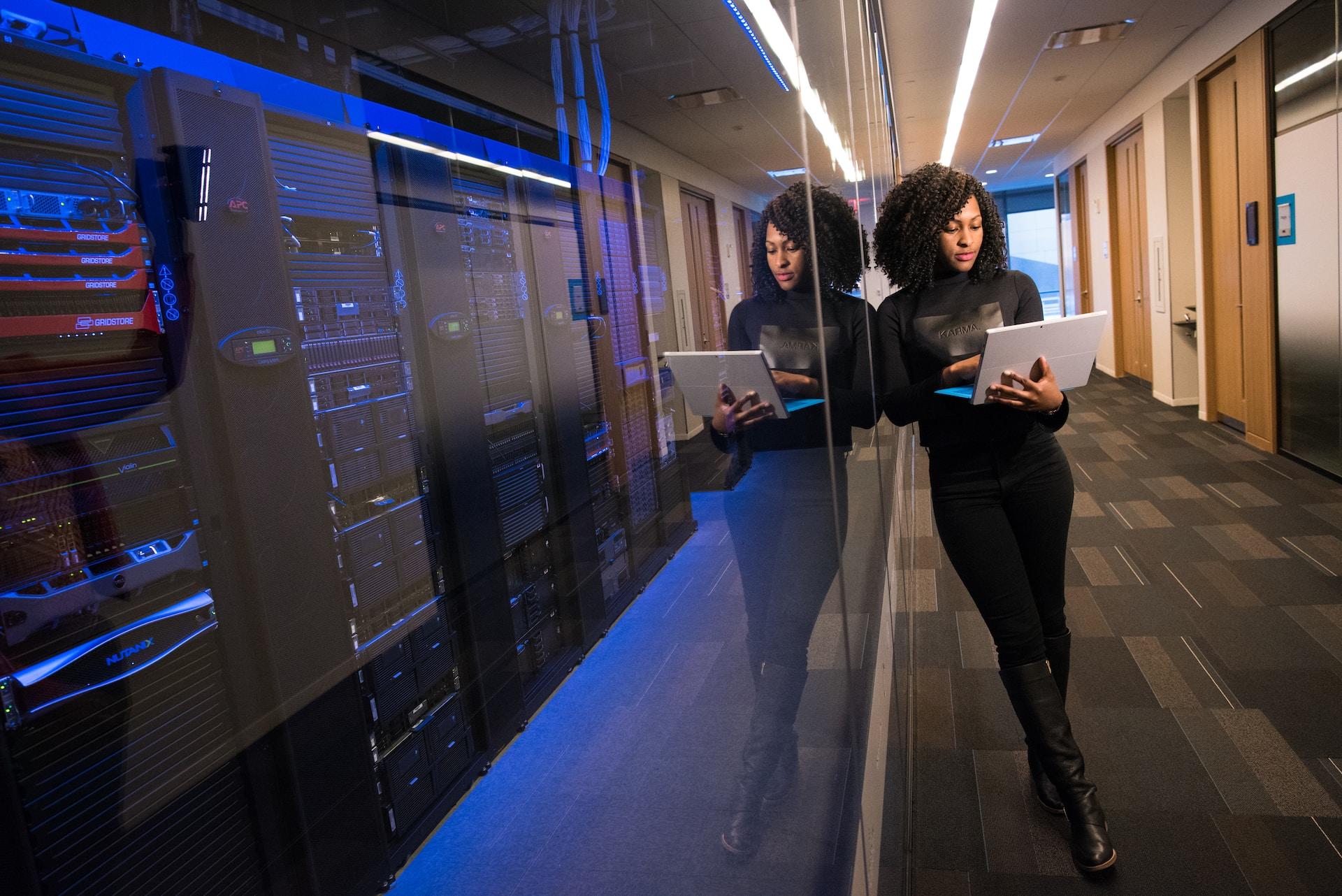 A person dressed all in black stands in an empty hallway, leaning against a glass wall behind which is a row of computer servers. 