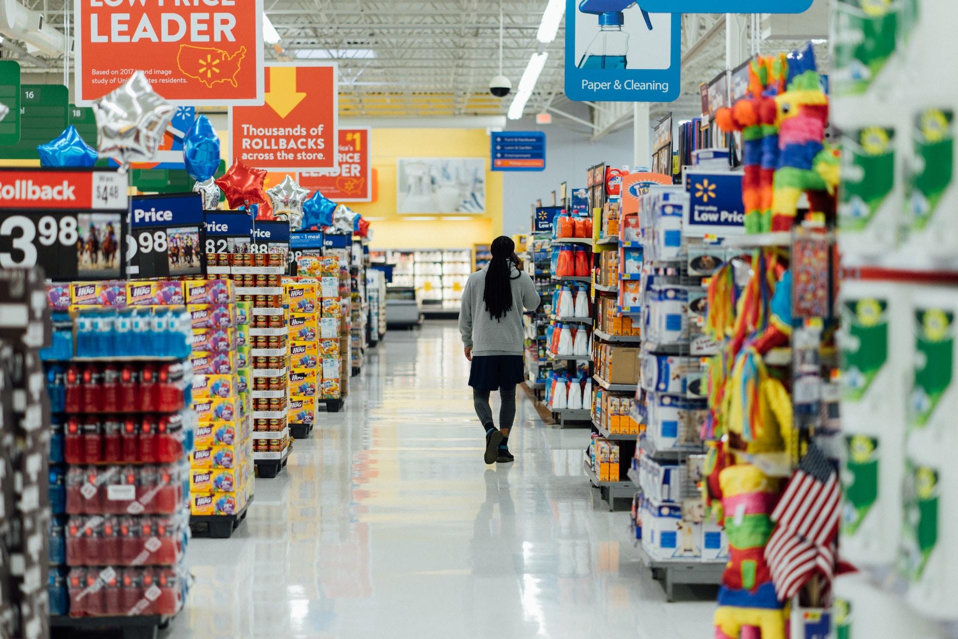 A person wearing grey and black walks down the aisle of brightly lit supermarket where colourful wares are on display and large orange signs hang overhead.