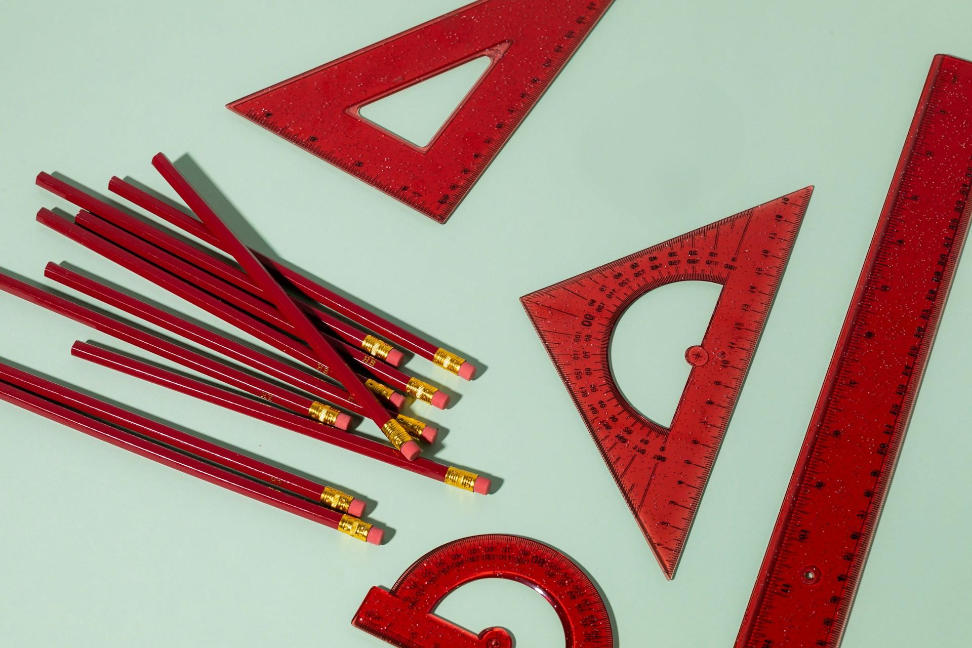 A collection of red pencils arranged alongside several red rulers on a neutral background.