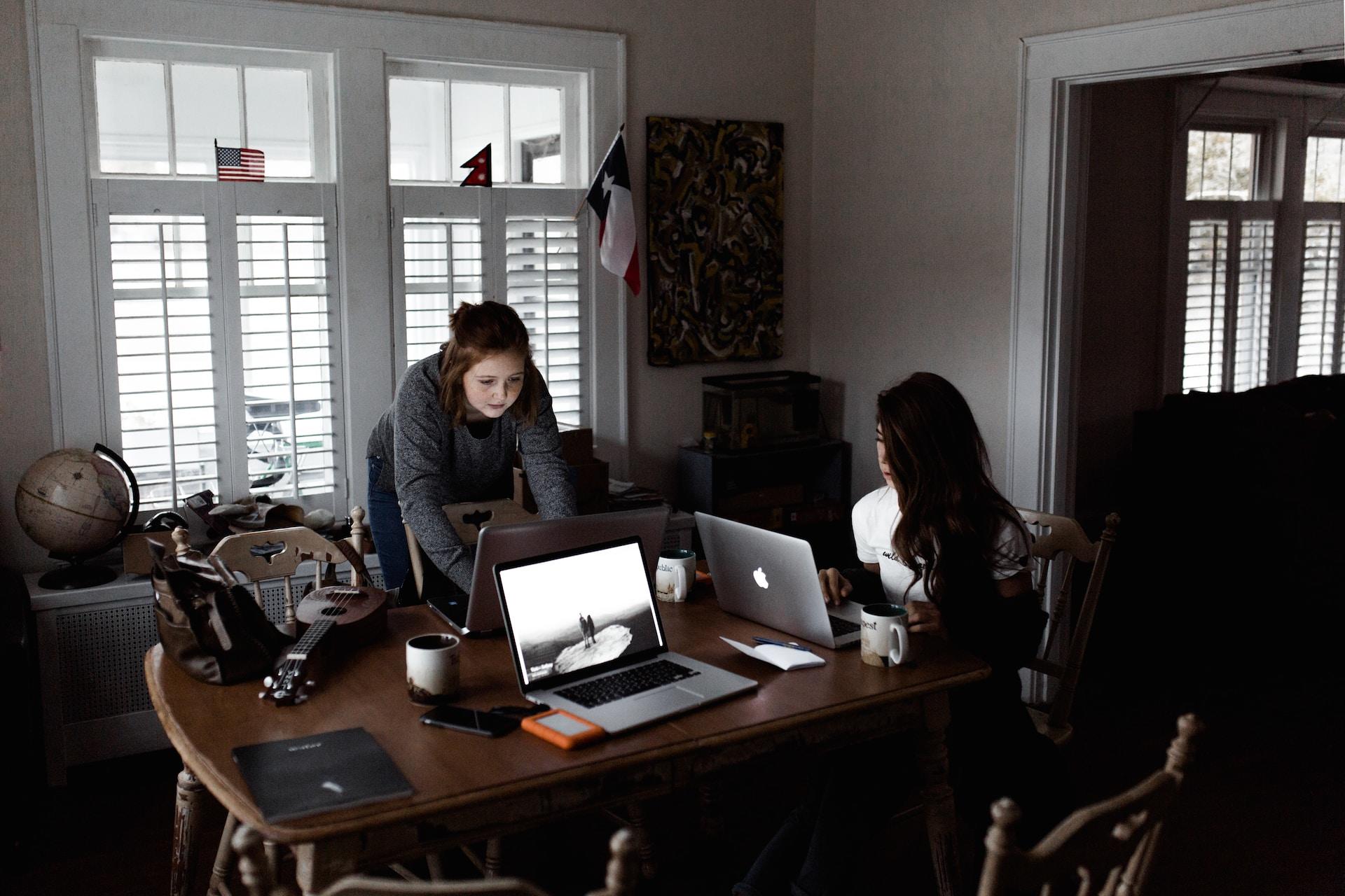 Two people work on their laptop computers, at a rectangular table that has three computers on it, in a room beset with shadows. 