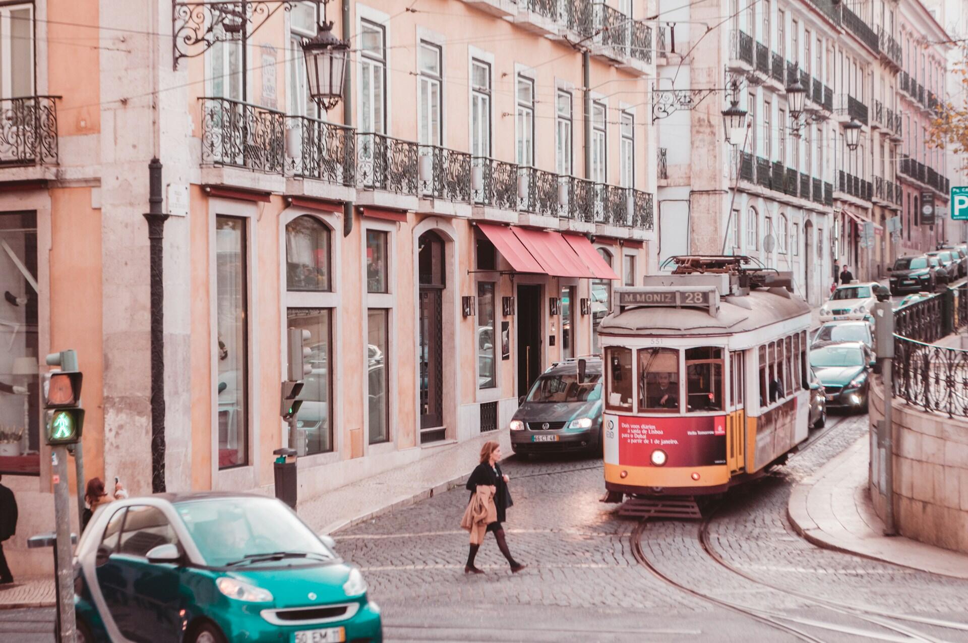 A vintage tram navigates a cobblestone street lined with shops and cars, under a soft, pastel-hued sky.