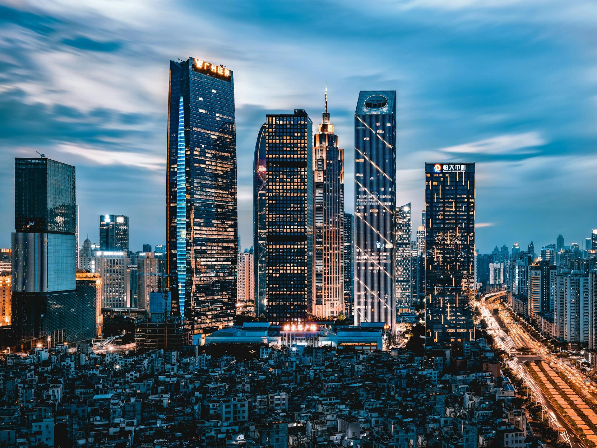 A city's skyline at dusk, with high-rise buildings discreetly lit up and a darker residential area in front of it and a well-lit highway alongside both.
