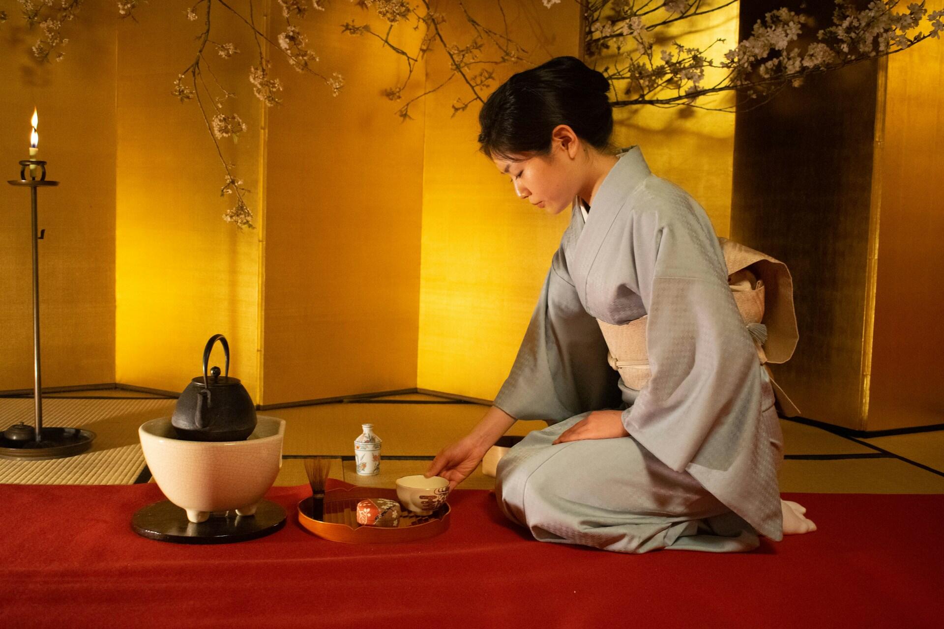 A Japanese woman wearing kimono and kneeling on the ground, preparing matcha in the traditional way