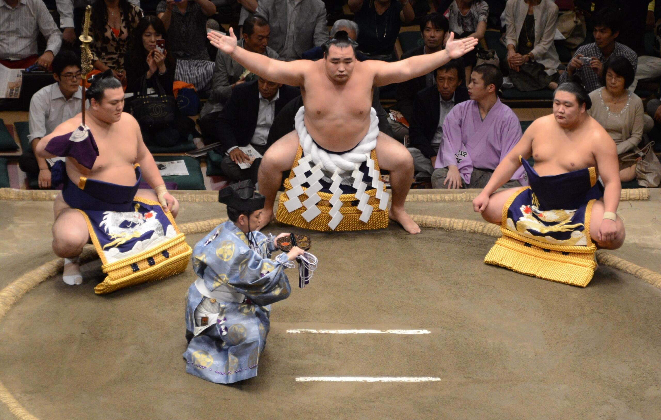 a yokozuna wrester wearing the iconic white rope belt with "lightning bolt" streamers with two sumo wearing silk aprons on either side in the ring