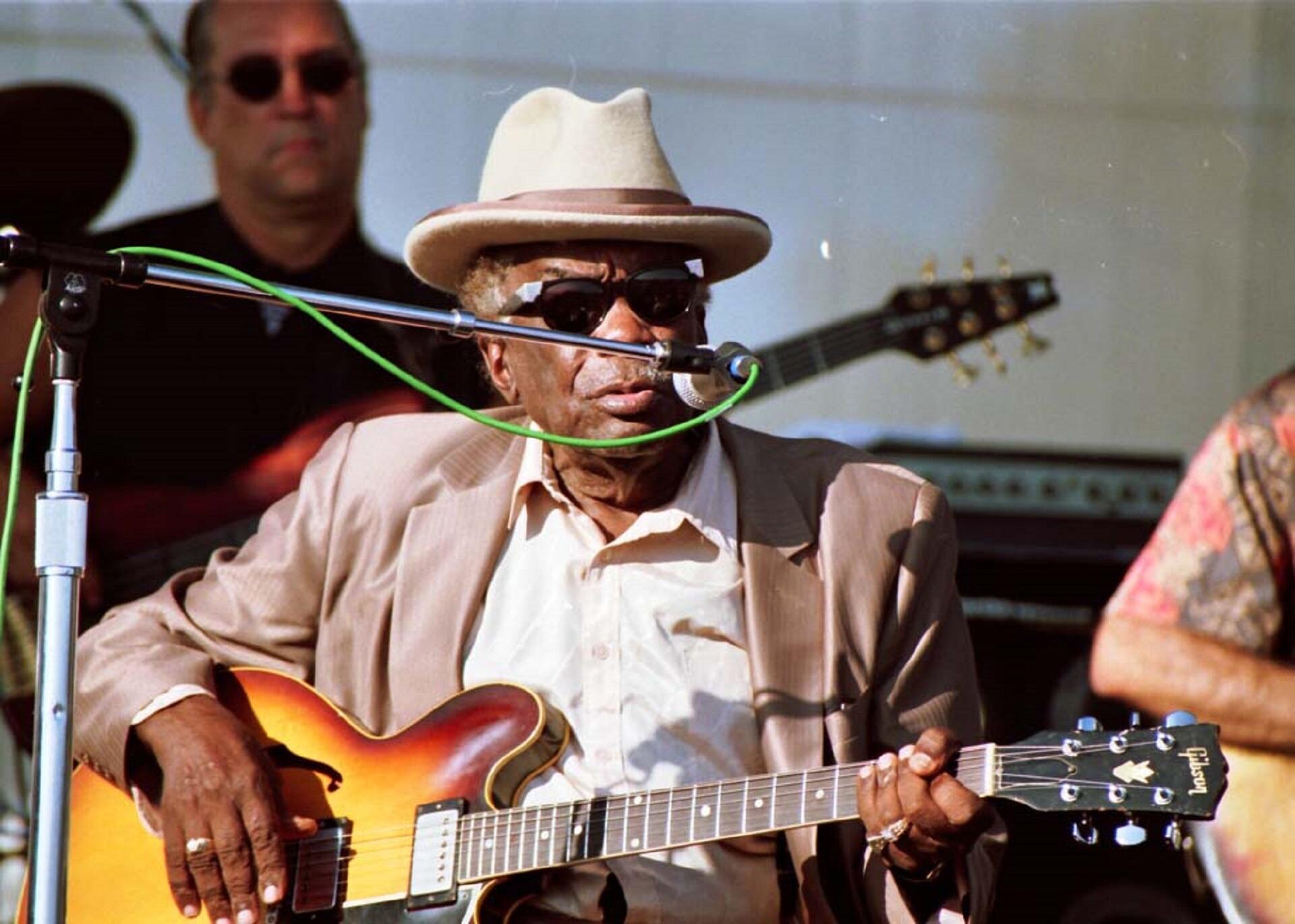 An older man wearing a tan hat with a brown brim and sunglasses holds a guitar while seated on a stage outdoors.