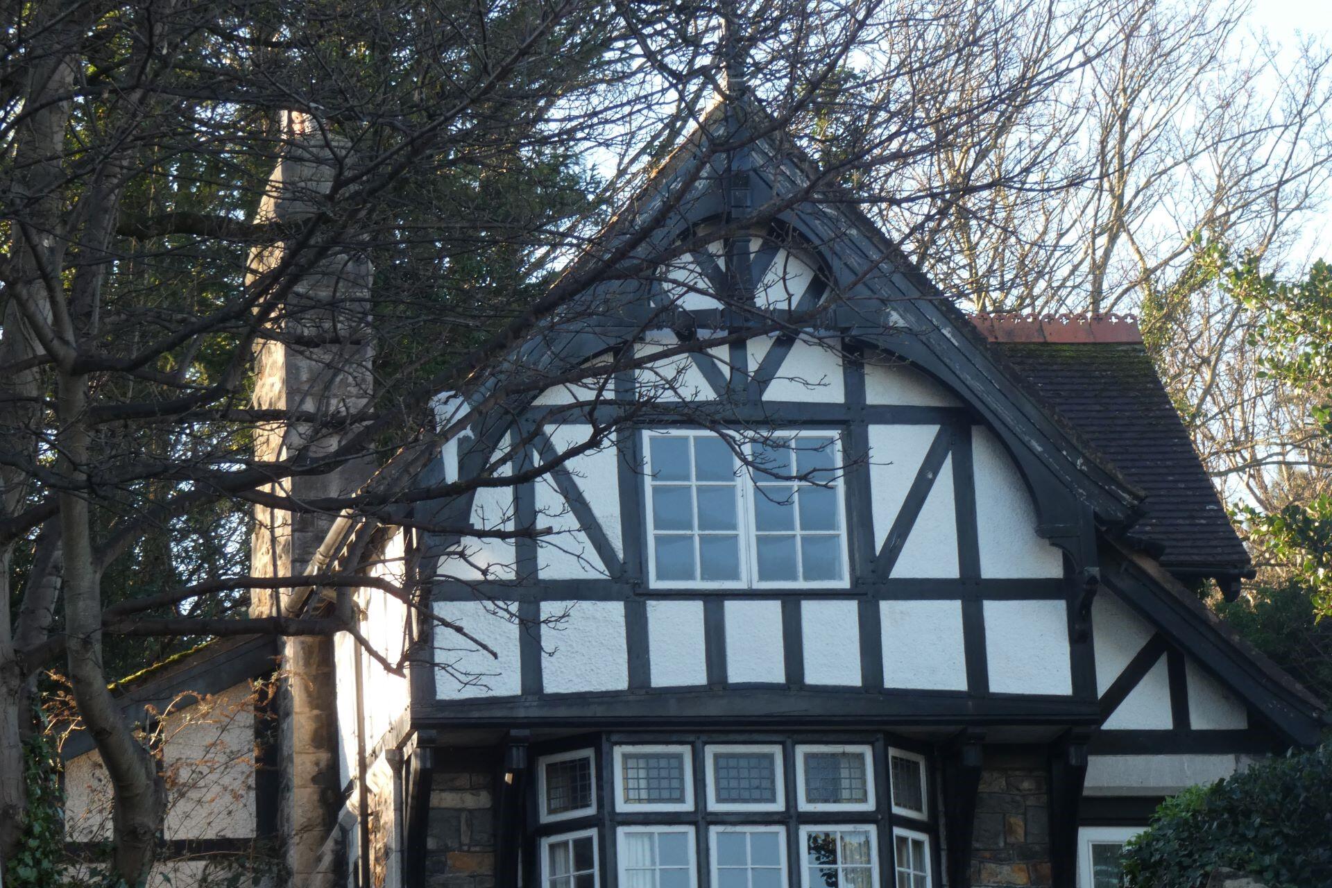 Black and white timber-framed Tudor-style house with a steep roof, multiple windows, and trees surrounding the building.
