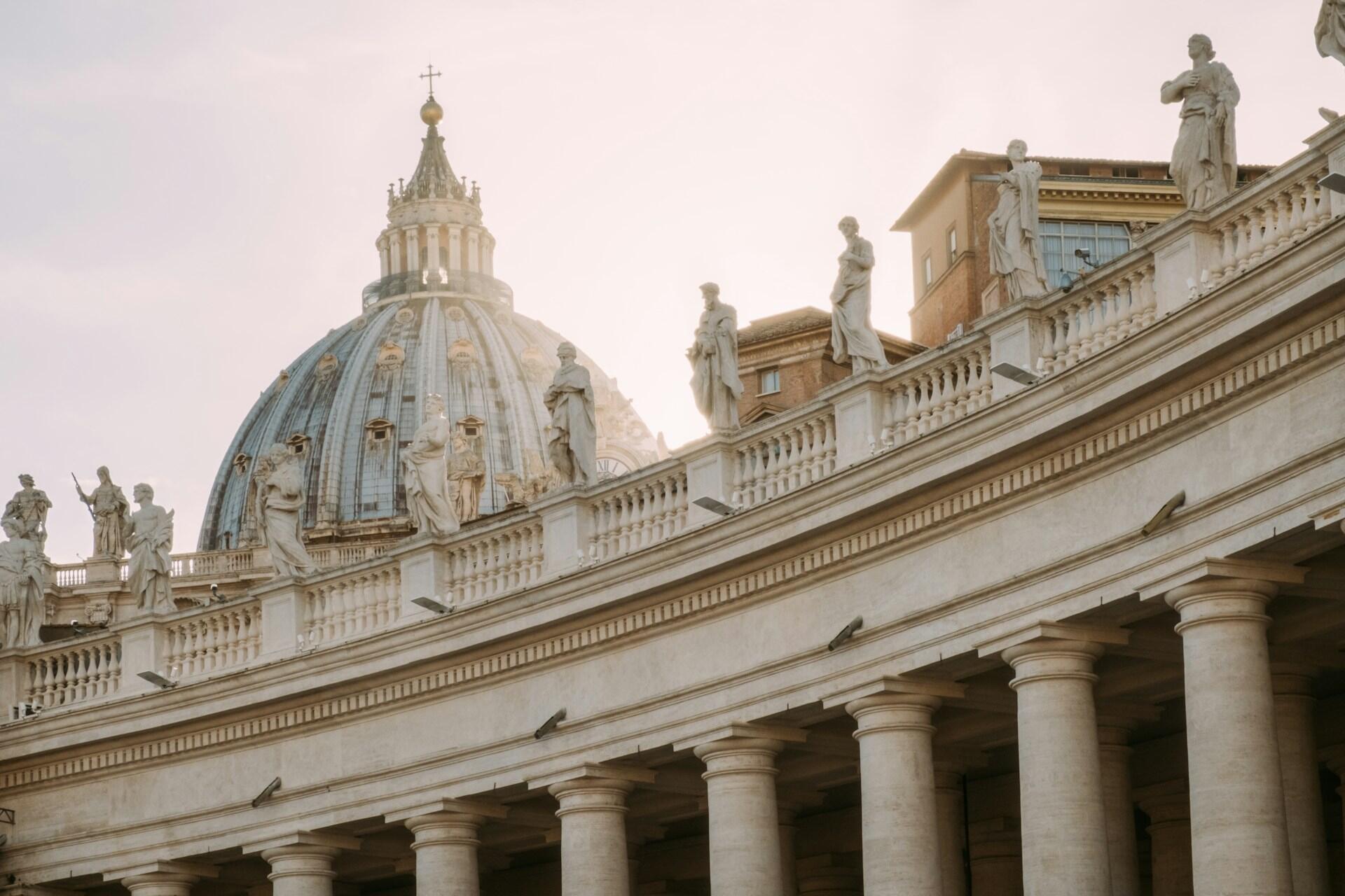 St. Peter's Basilica dome captured against a pastel sky, with statues lining the balustrade and classic columns in the foreground.