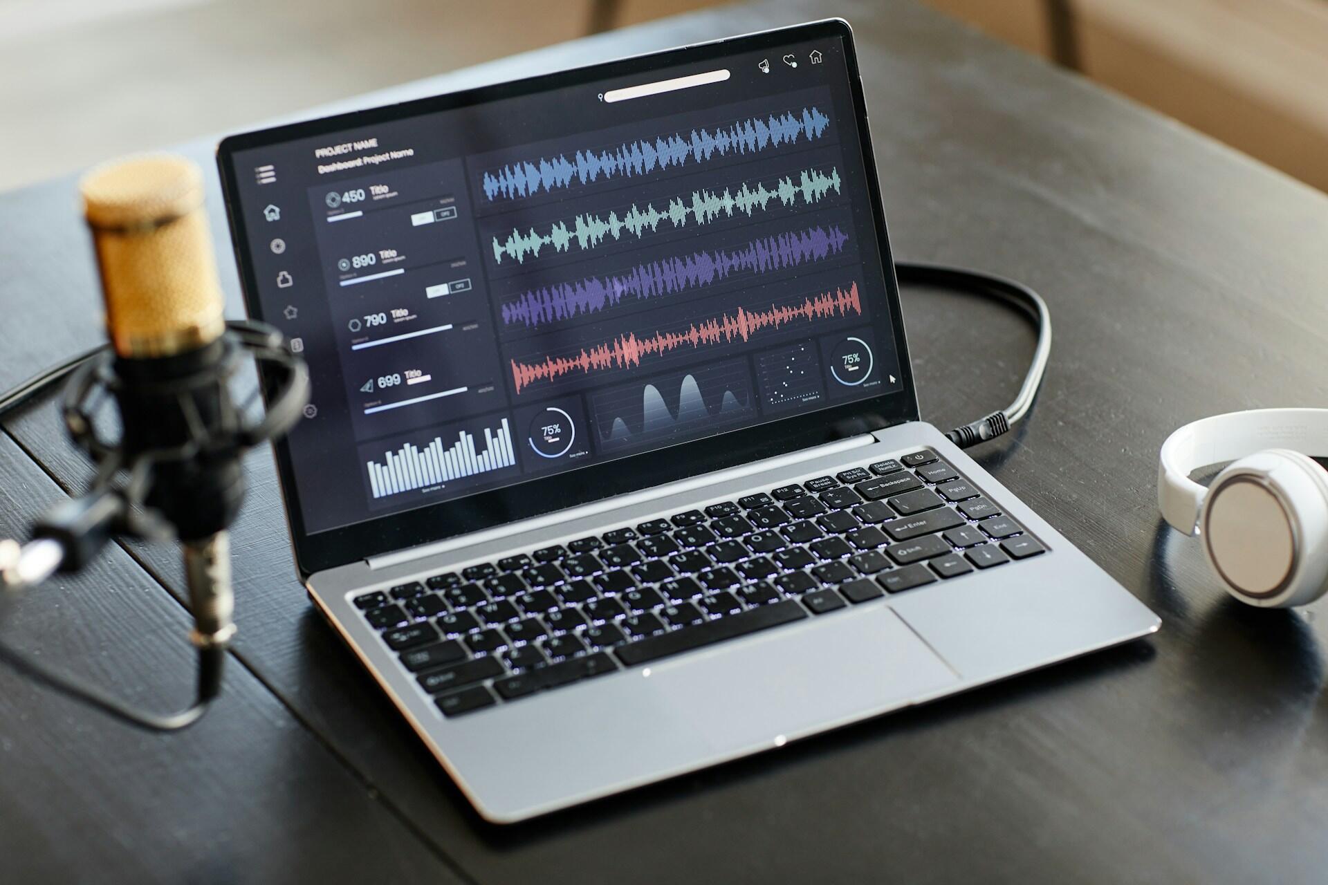 A laptop on a wooden table displaying audio waveforms, with a microphone and headphones nearby.