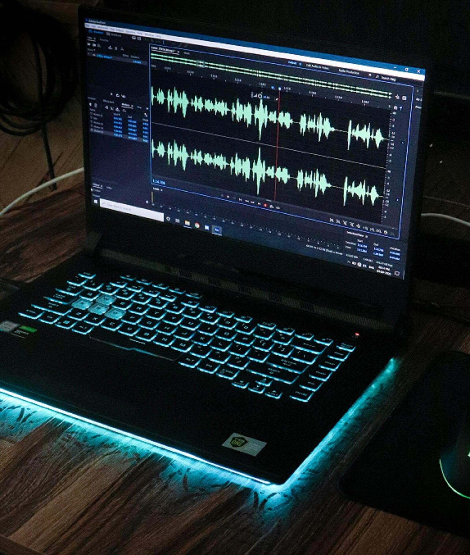 A laptop with teal backlit keys displays audio editing software. Visible soundwaves on the screen suggest a focus on sound production.