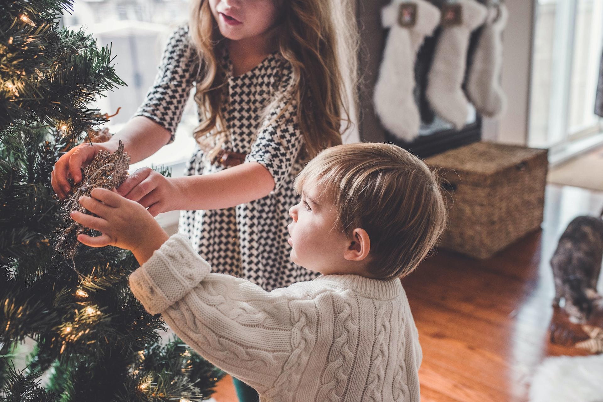 Two children hanging ornaments on a Christmas tree with white Christmas stockings hung on the wall in the background.