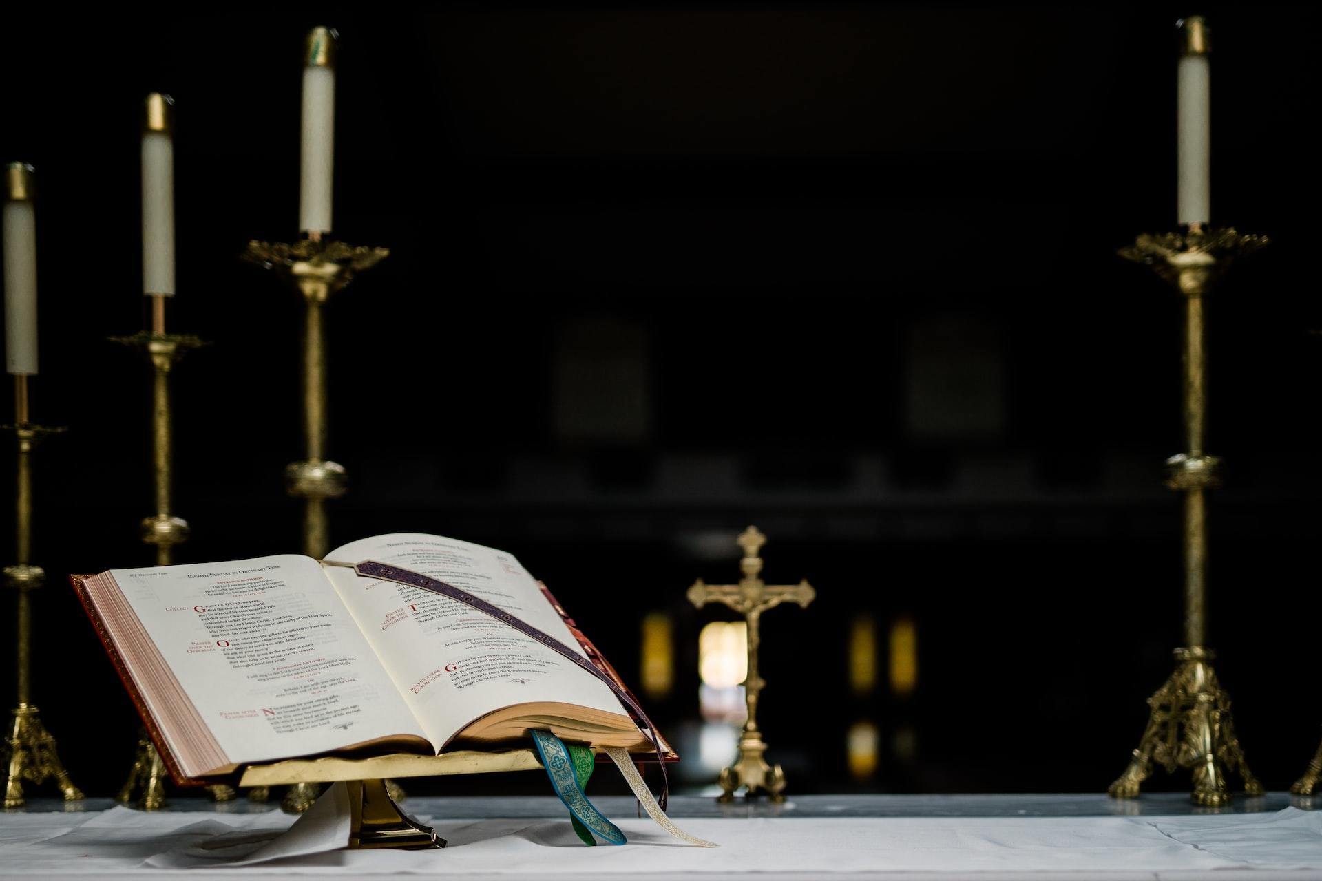 An cross, several candlesticks and an open hymnal resting on a stand all sit atop a white-cloth-covered altar against a black background.