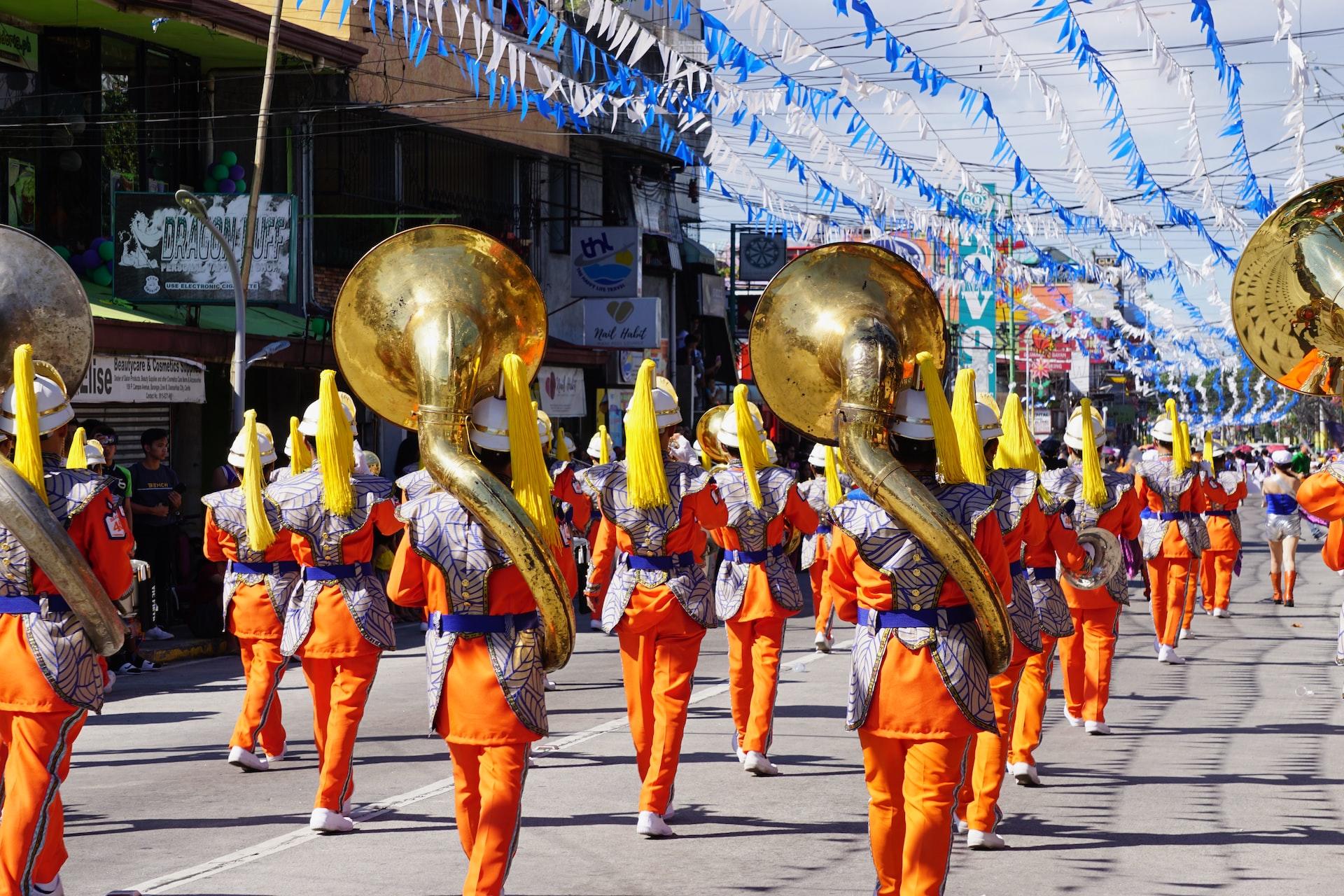 A marching band viewed from the back, bedecked in orange and silver uniforms, carrying trombones and other instruments on a sunny day.