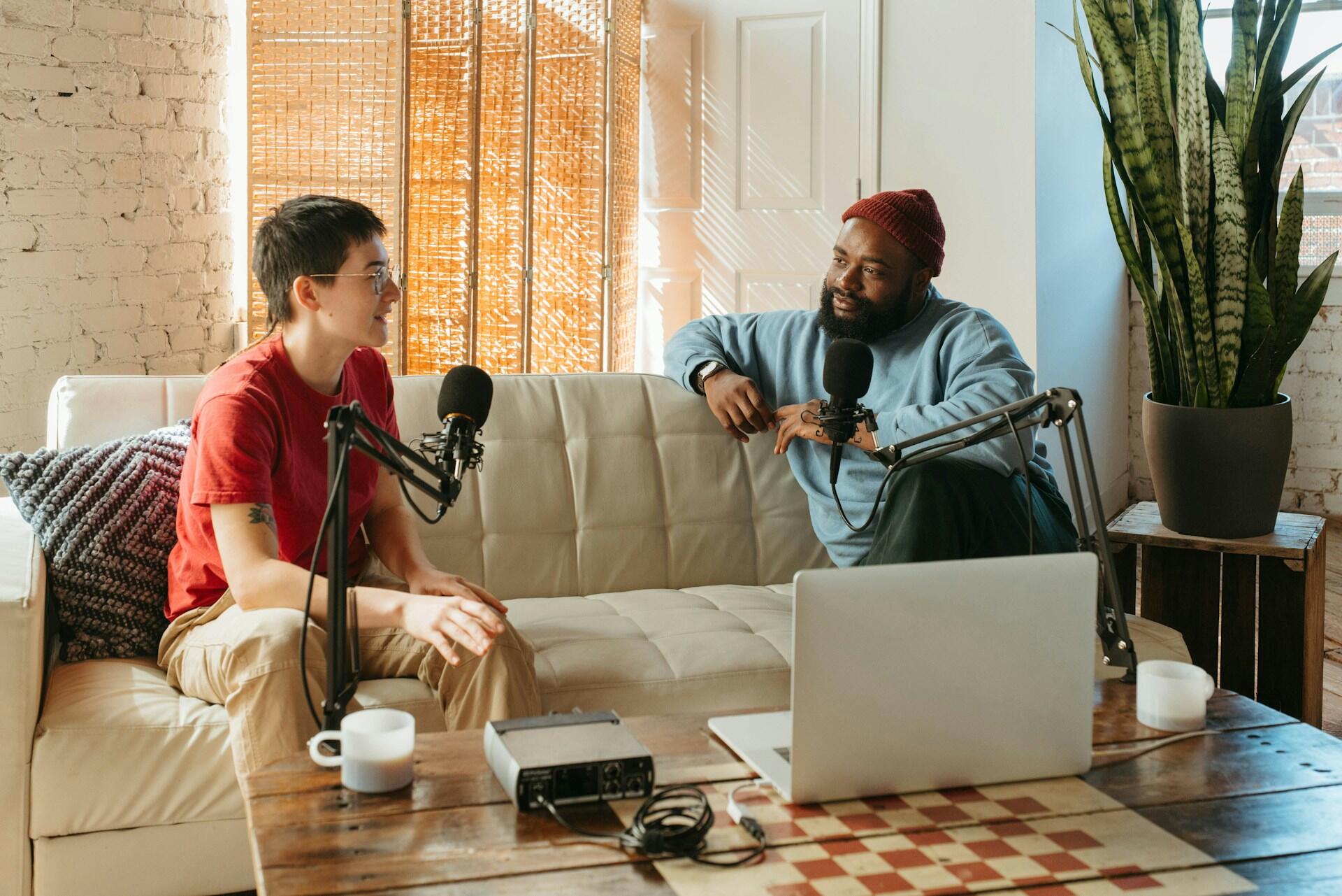 Two people sit on a couch with microphones, engaged in conversation. A laptop and audio equipment are on a wooden table nearby.