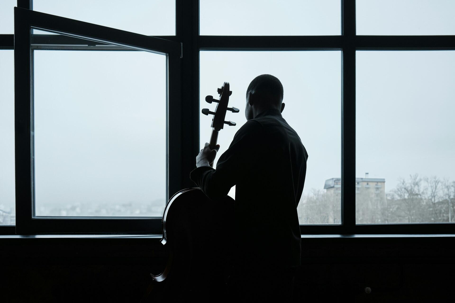 A silhouette of a man holding a cello by an open window, gazing out at a cloudy urban landscape.