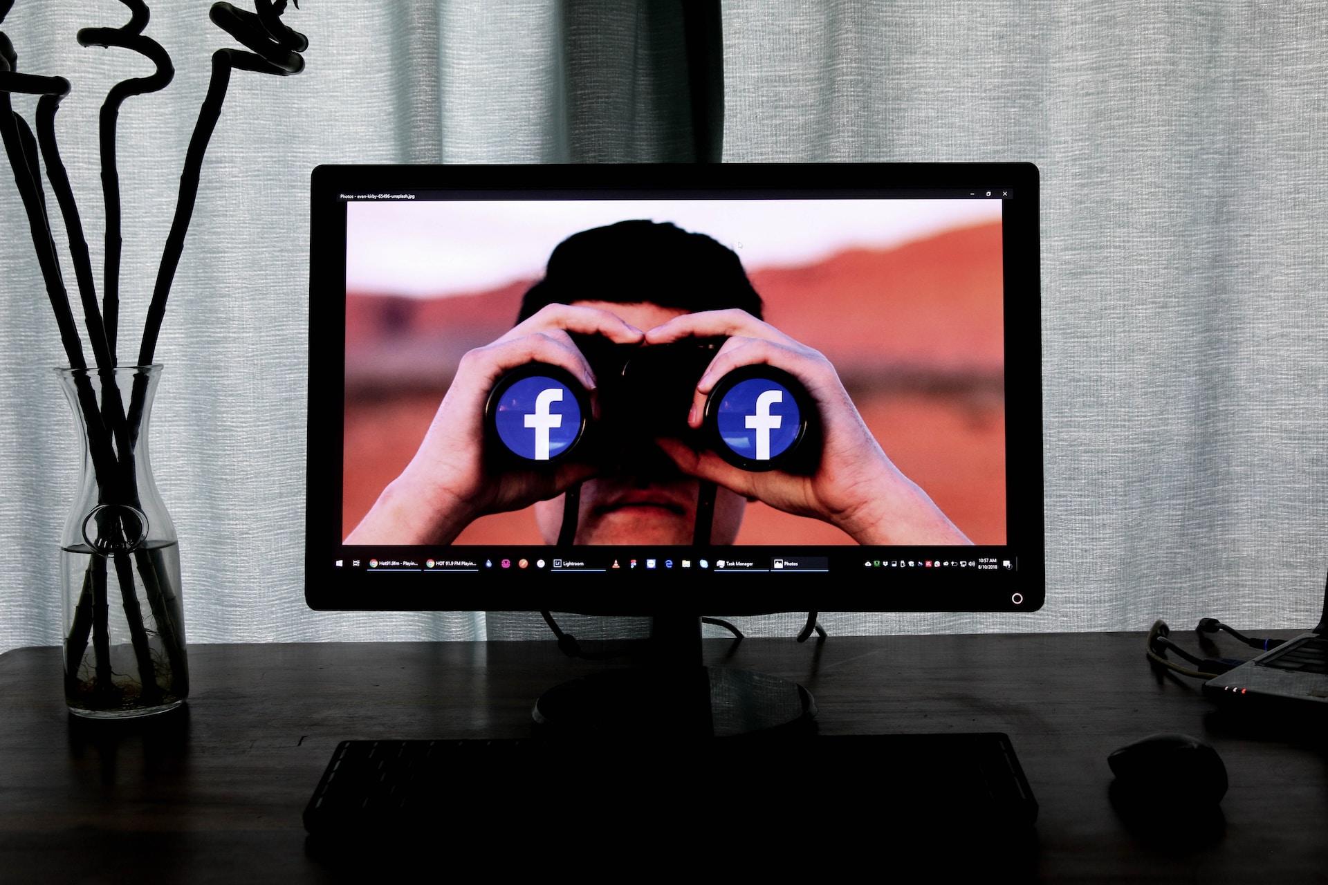 A television screen set on a black surface next to a vase containing three sprigs of lucky bamboo, displaying a person holding binoculars to their eyes which display the facebook logo, set against an ochre red background.