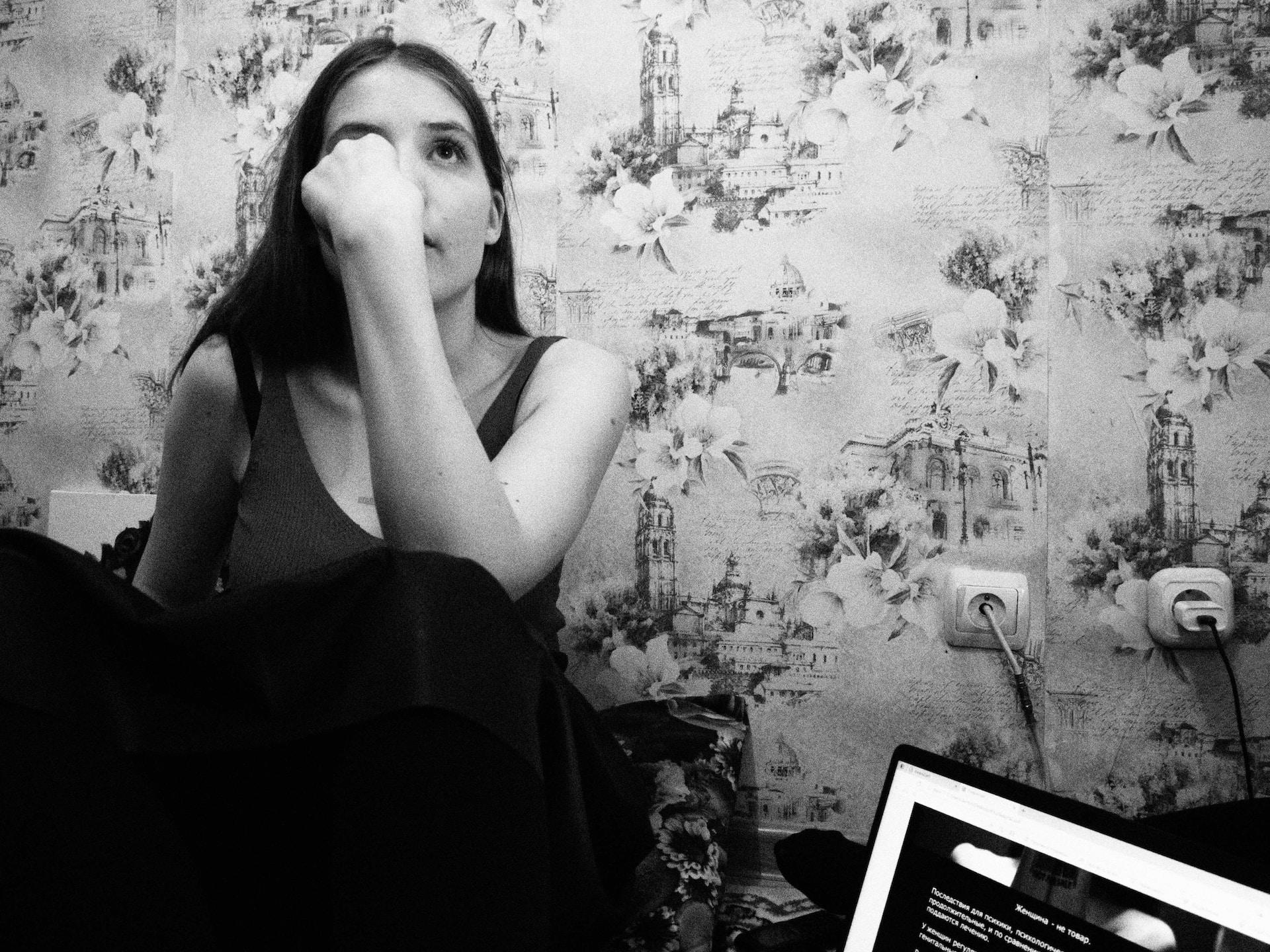 A greyscale image of a young woman wearing a tank top propped up against a floral patterned wall with an open laptop computer close by, wiping her eyes.