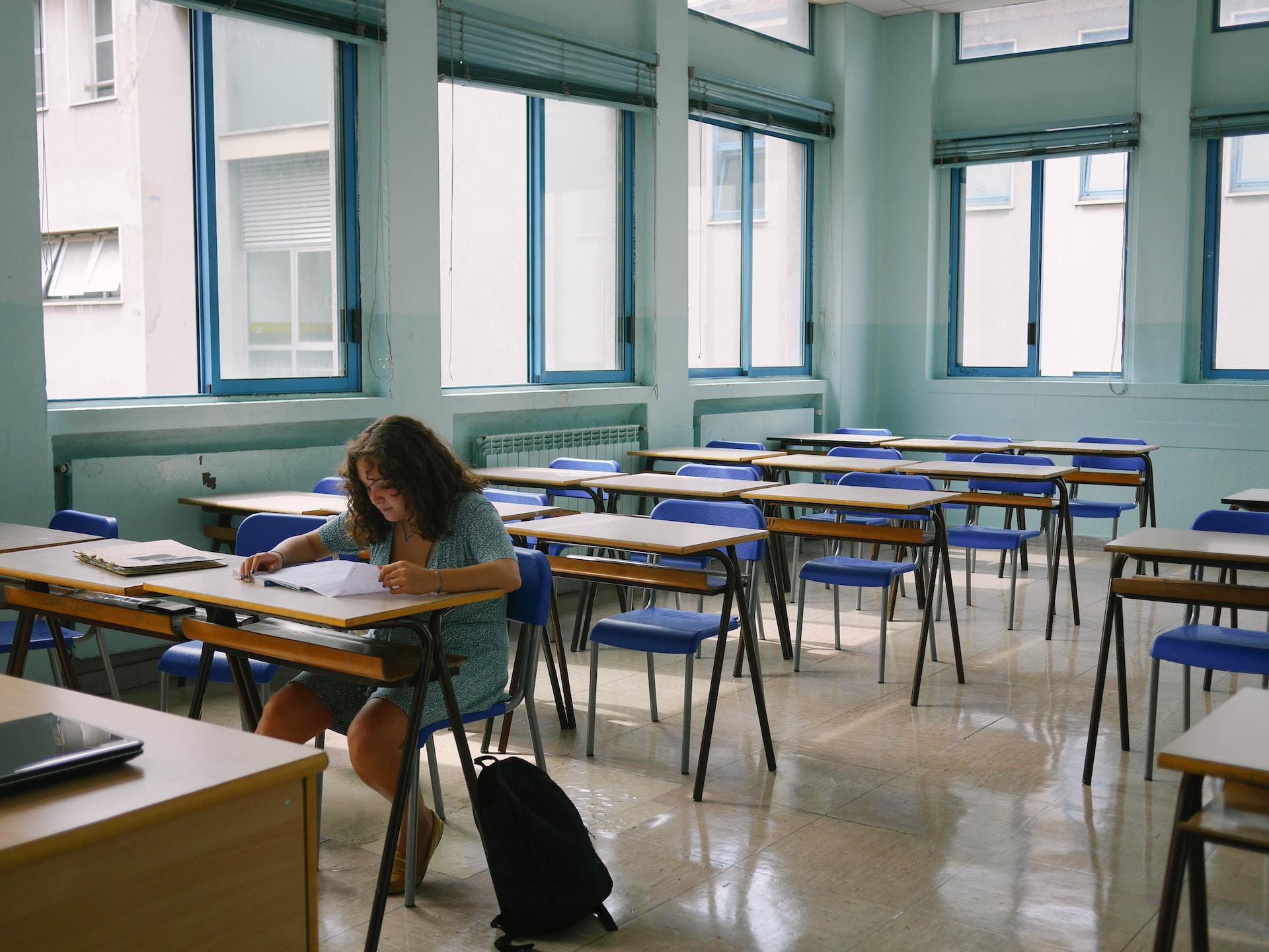A person sits alone in a classroom full of wooden desks and blue chairs with light streaming in from the room's many windows.