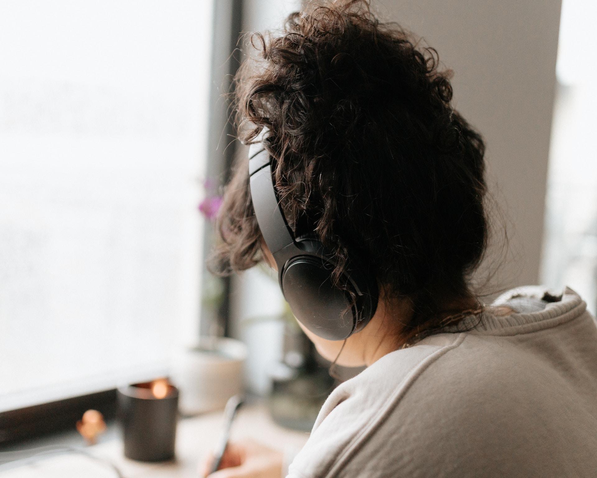 A person wearing a light coloured shirt wears black headphones as they sit at their desk with a pen in their right hand.