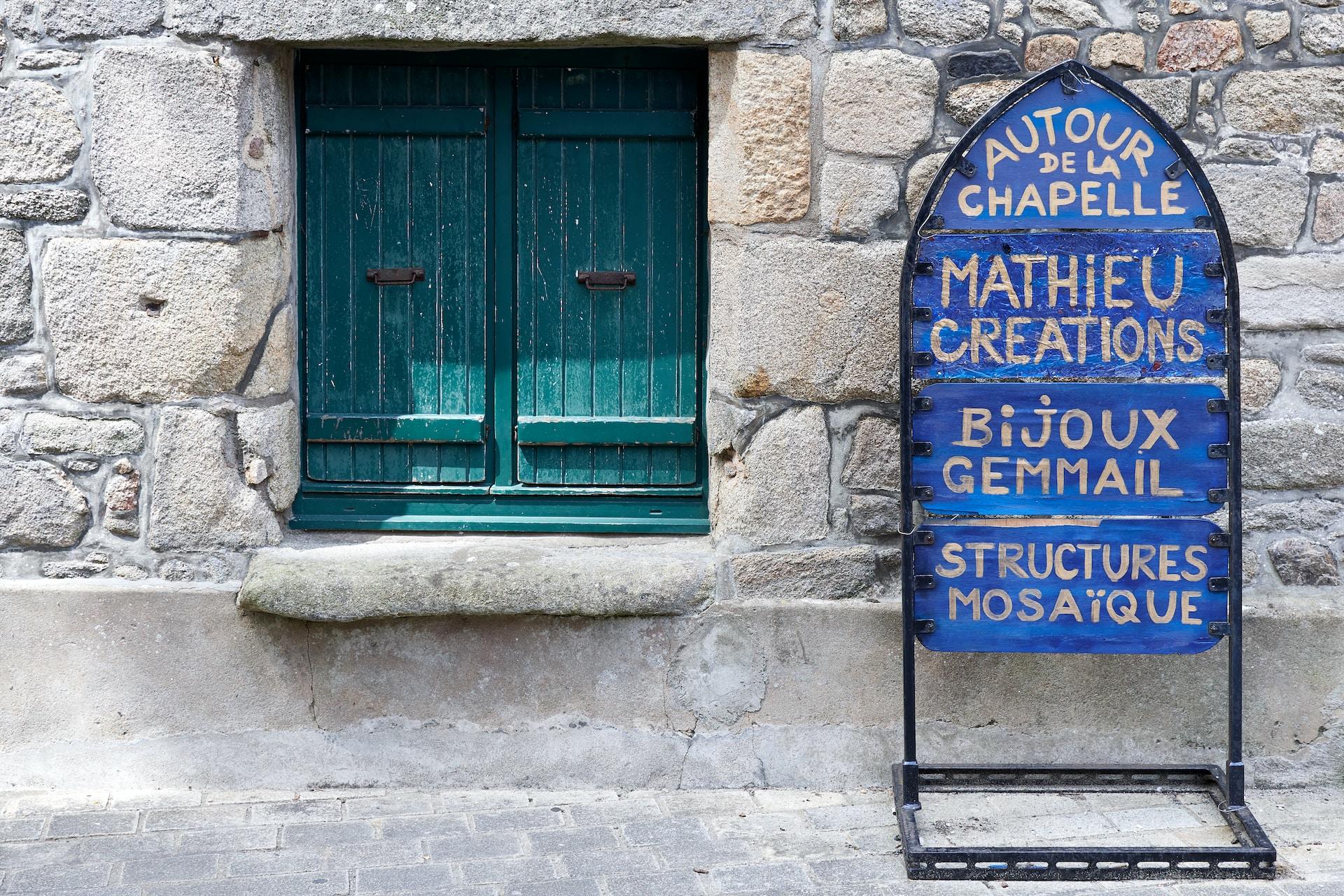 A blue sign with French writing in white encased in a narrow black frame next to a green door set into a white stone wall. 