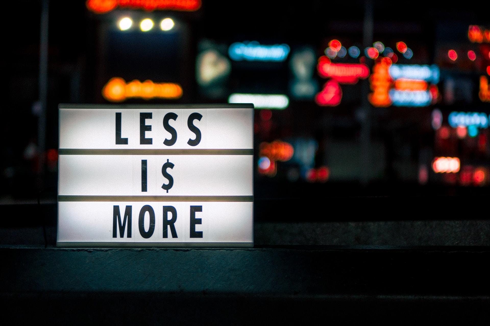 A lighted letter box with less is more spelt out on it against a nighttime background with hazy, colourful neon in the background.