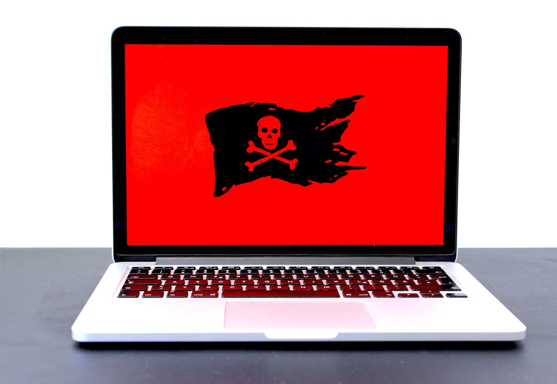 A white laptop computer with a black keyboard sitting on a grey surface, displaying a red screen with a black skull and bones flag.