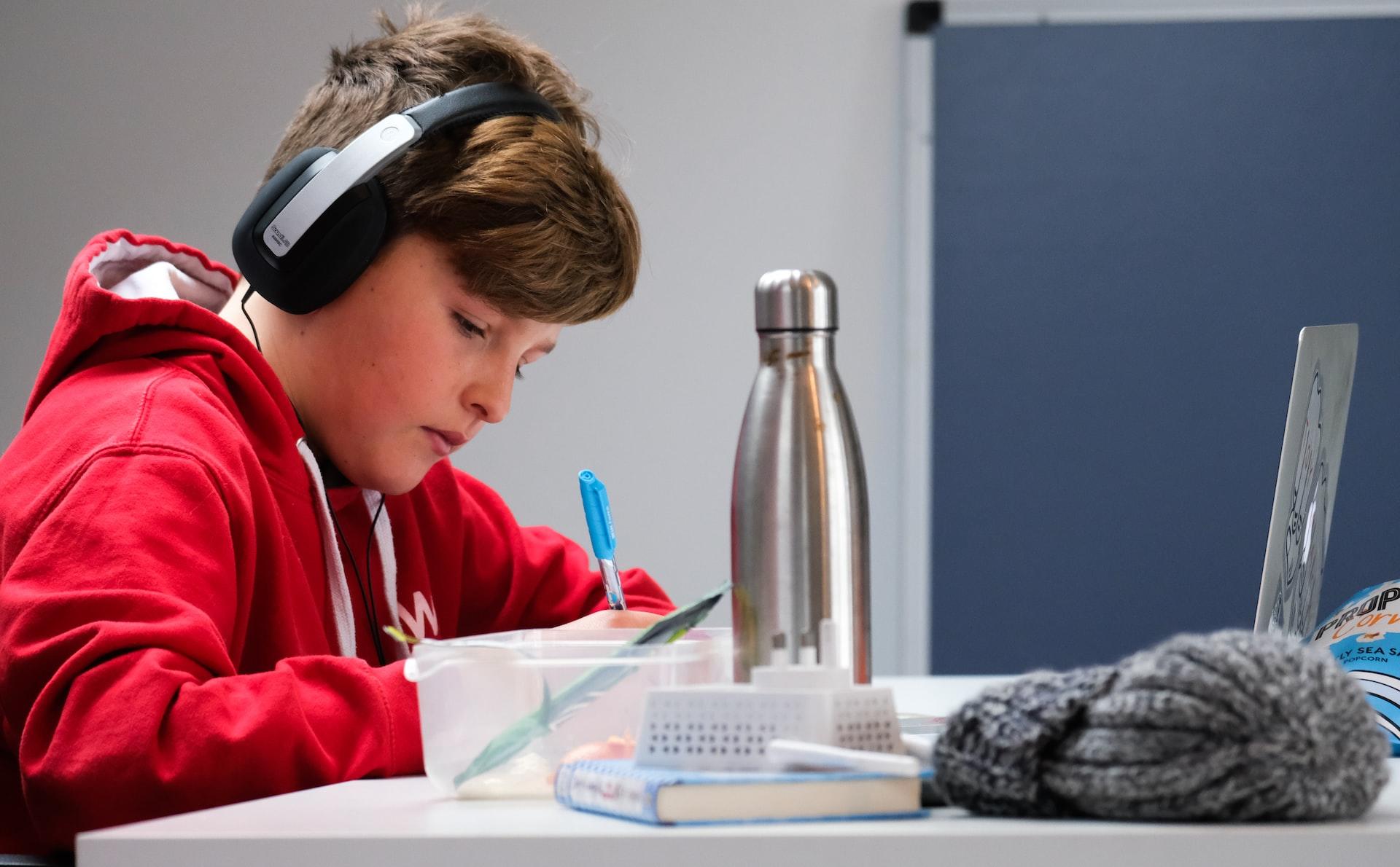 A boy wearing a red top and headphones over his ears sits and writes in front of an open laptop with a metal flask by his side.