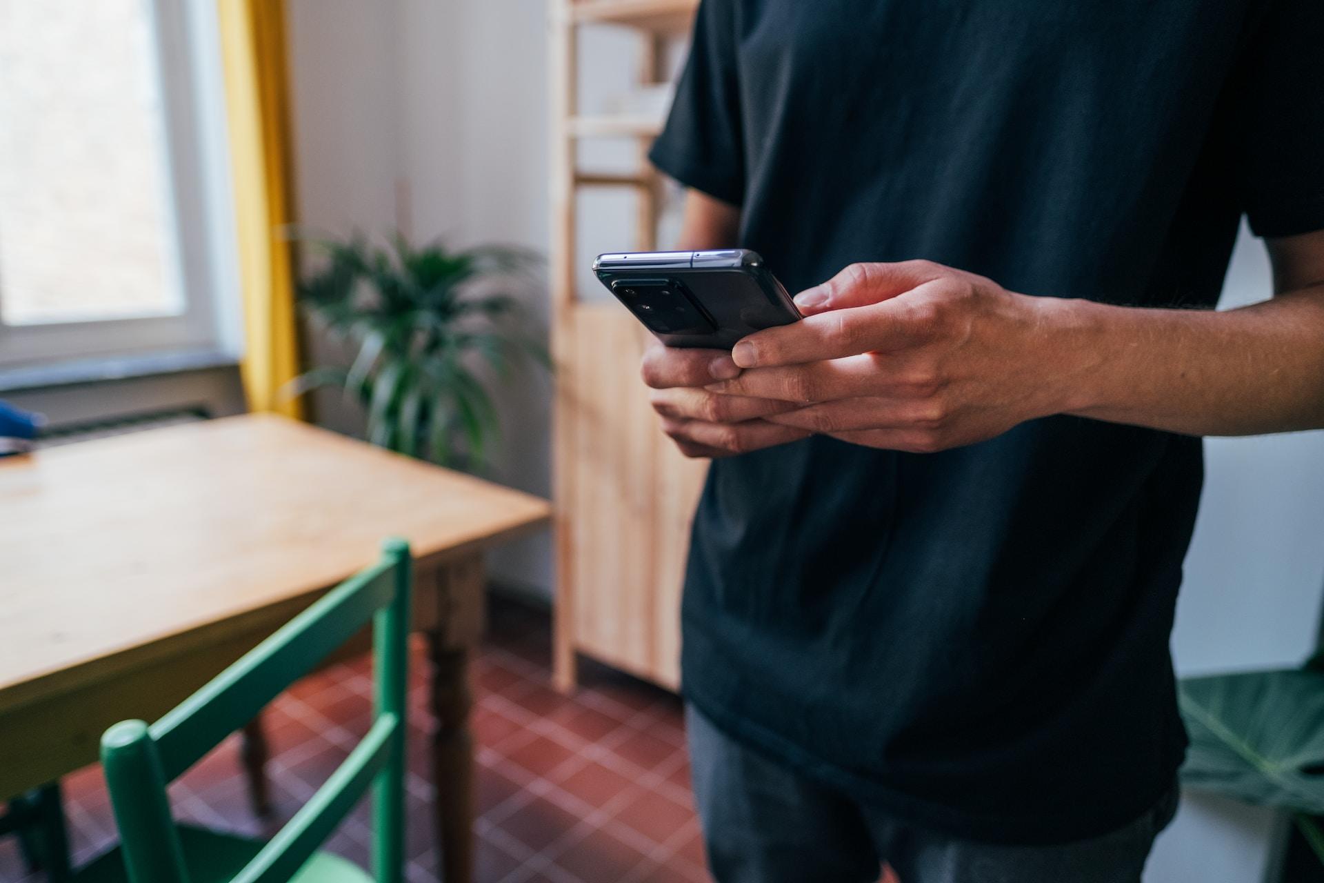 A person wearing dark coloured clothing stands next to a wooden table with a phone in their hands as they tap the device's screen.