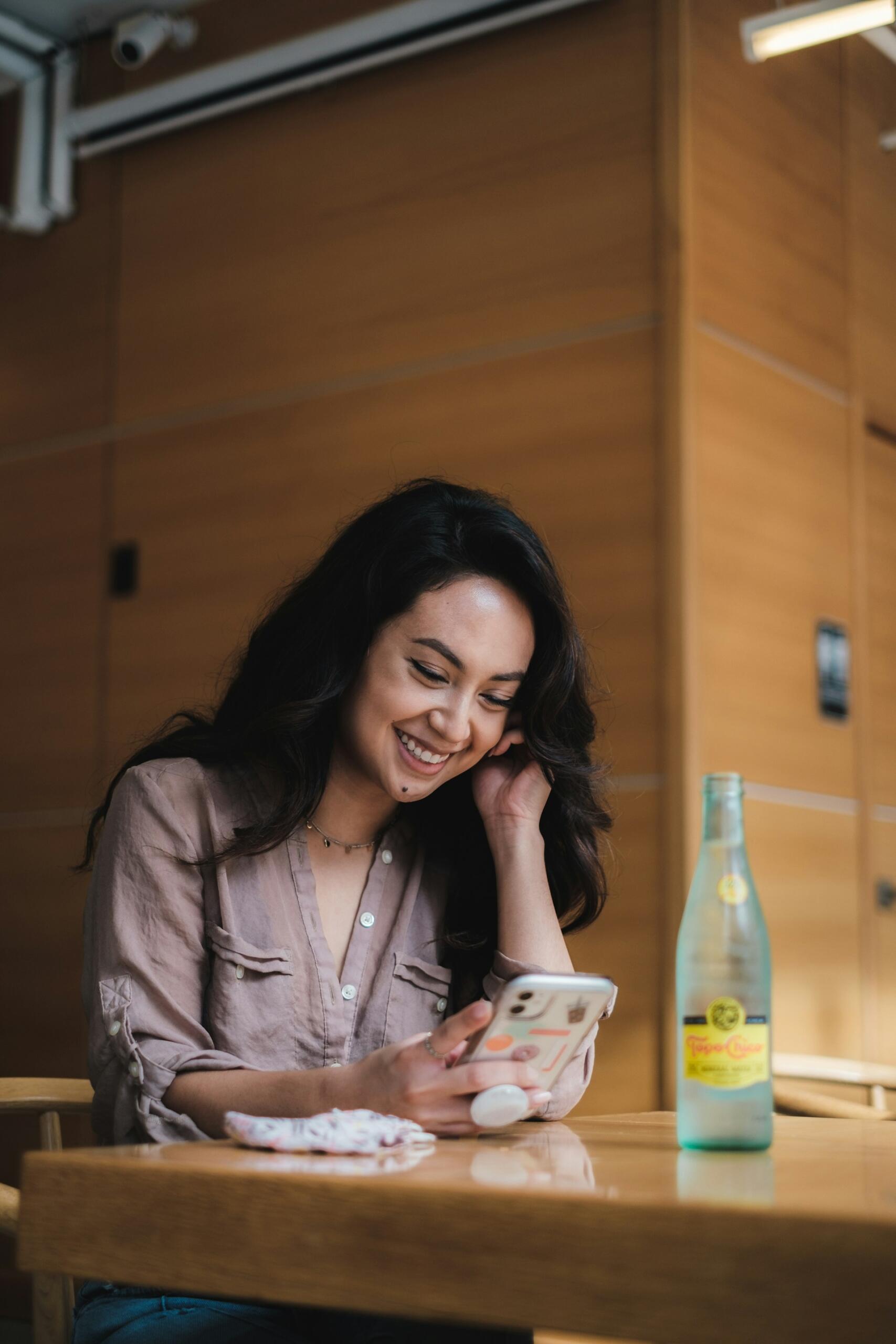 woman sitting at a table in a cafe, looking at her mobile phone and smiling
