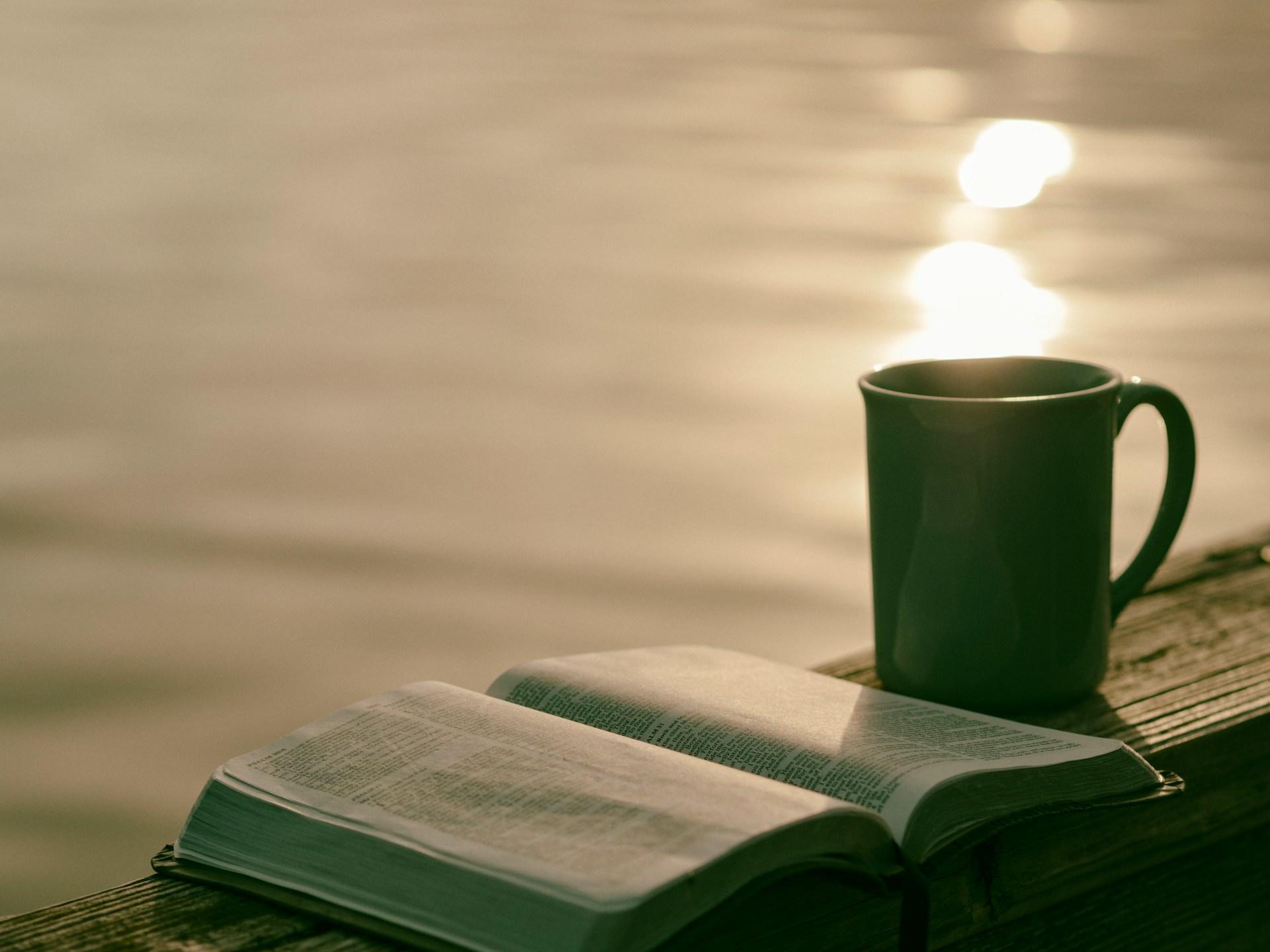 A cup of coffee rests beside an open book on a wooden bench overlooking a serene body of water.