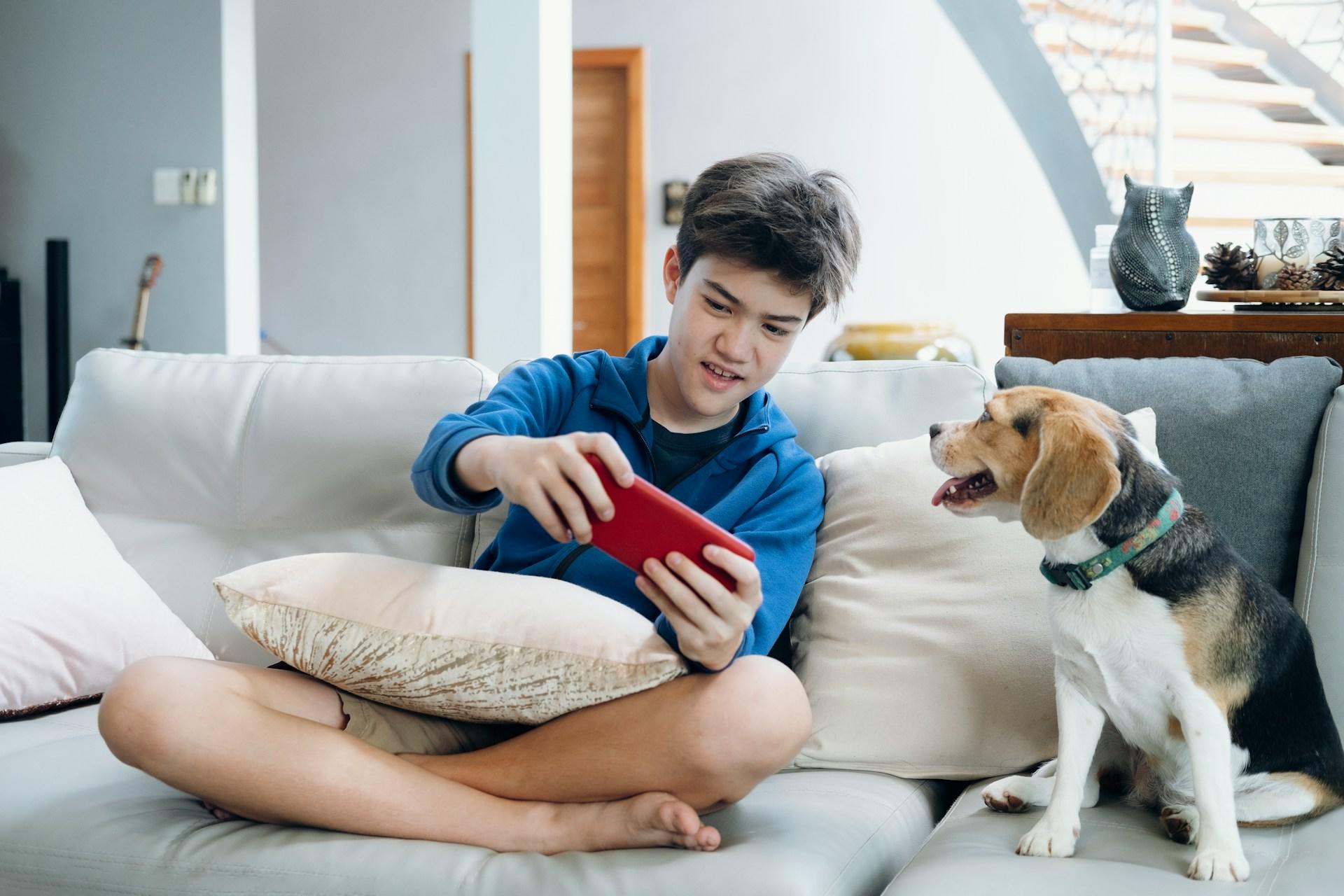 A youth wearing a blue top and short pants sits cross-legged on a white sofa, holding a phone in a red case so that the dog sitting next to them can see the screen.