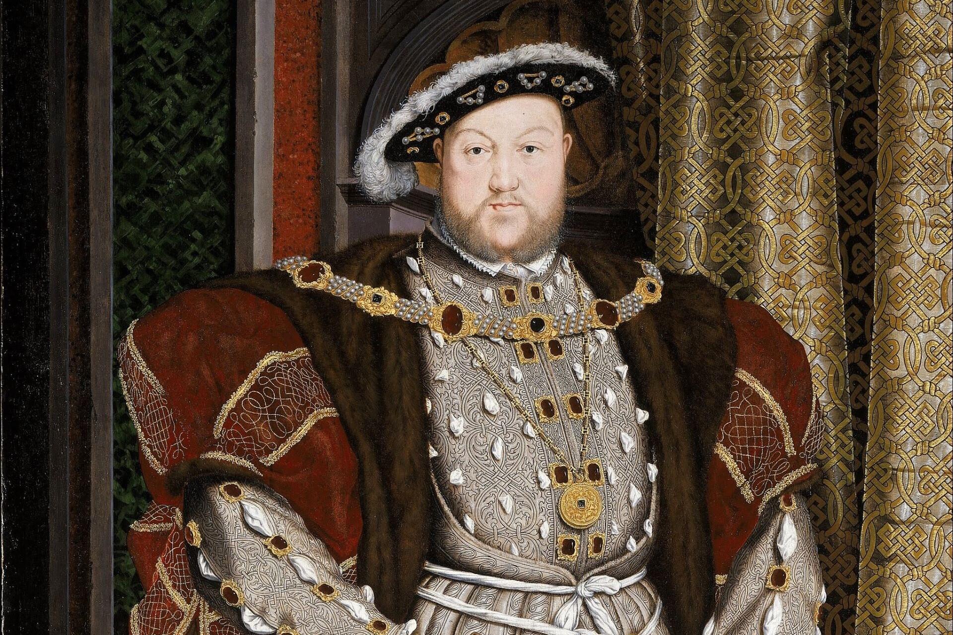 Portrait of a large man with a beard wearing ornate Tudor clothing decorated with jewels, embroidery, and fur trim, standing indoors beside patterned drapery.