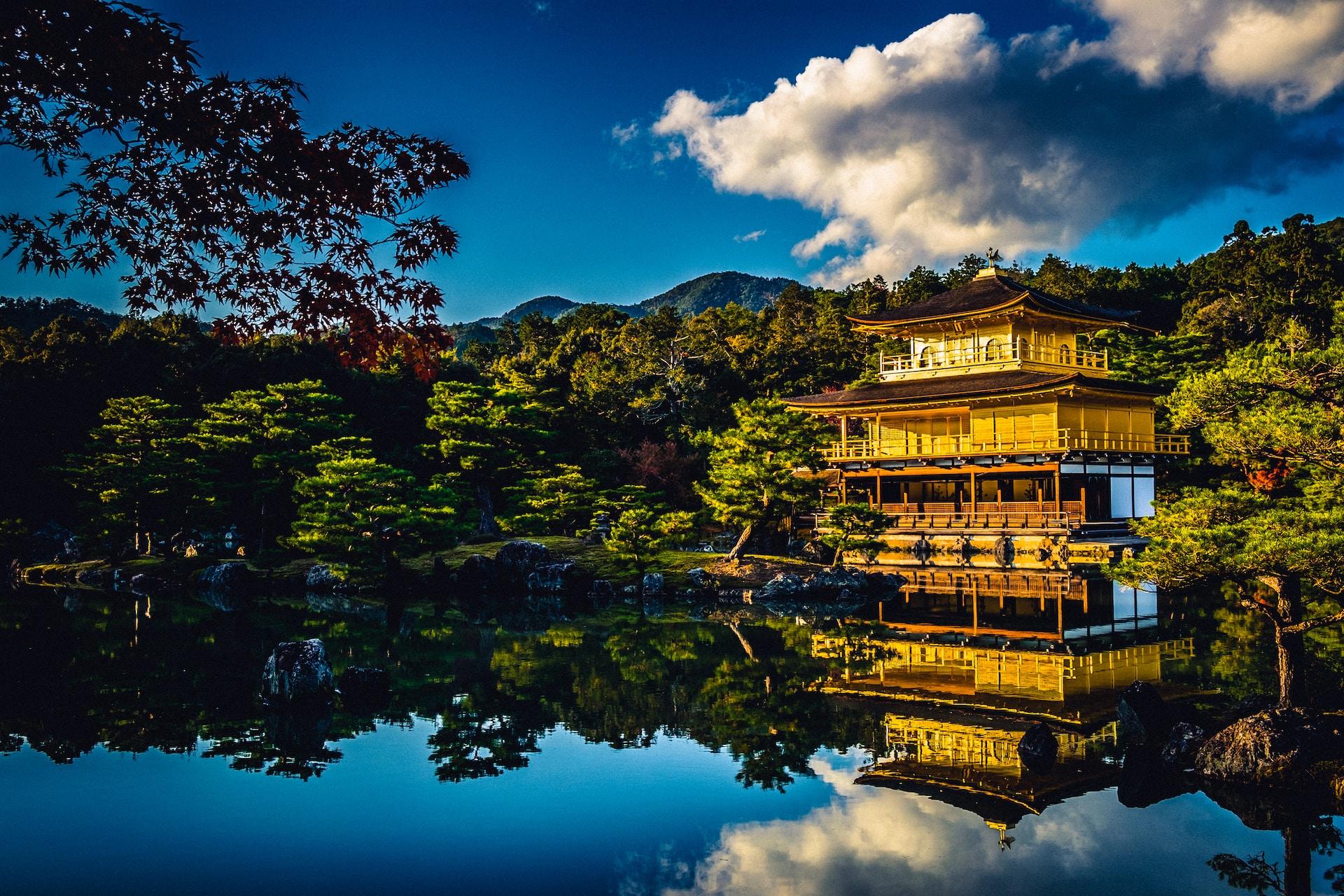 Pagoda House with its golden facade, reflected in the clear blue waters of the lake it fronts with a puffy cloud contrasting against the deep blue sky. 