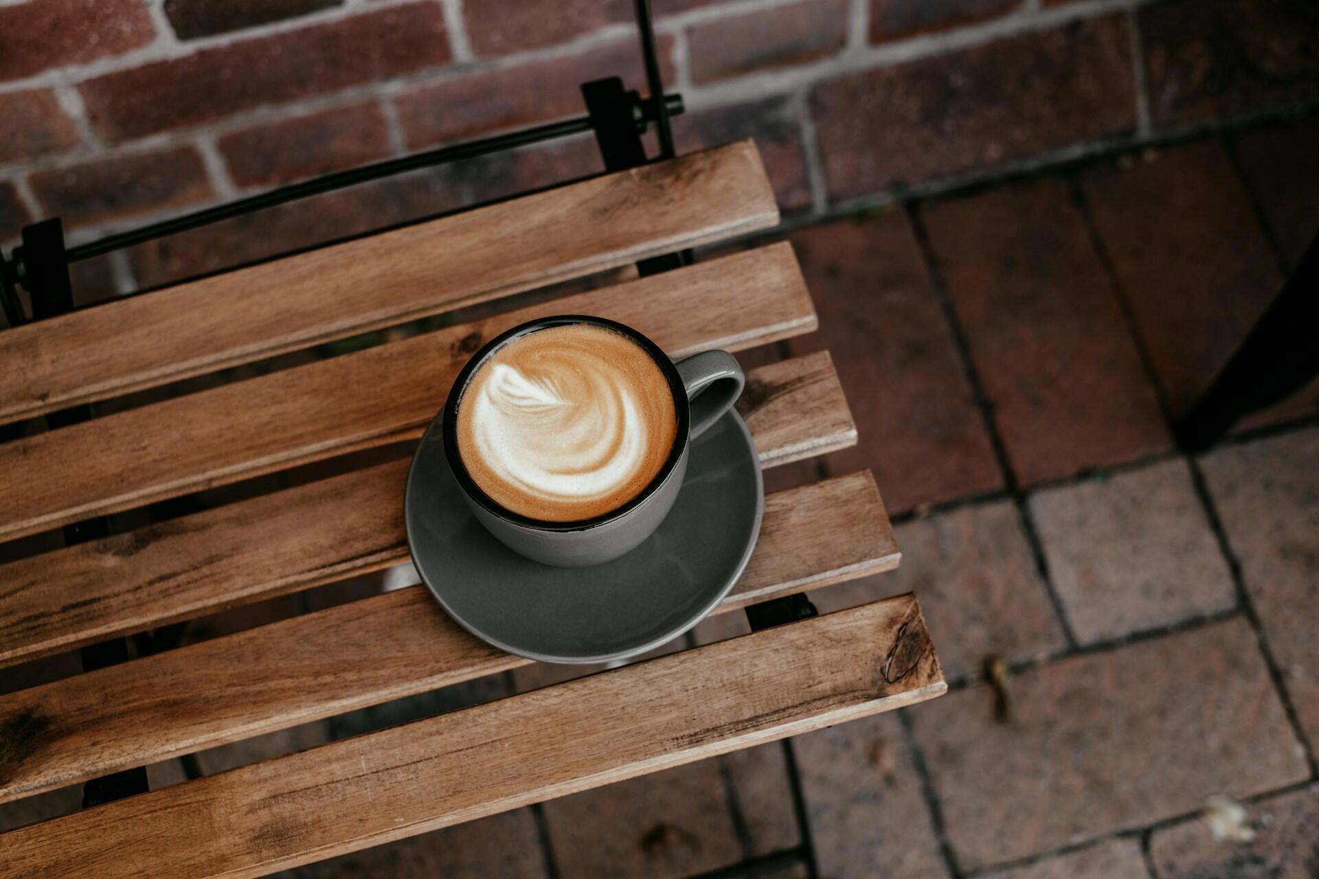 A coffee cup with frothy latte art sits on a round gray saucer atop a wooden table outdoors, with a brick wall in the background.