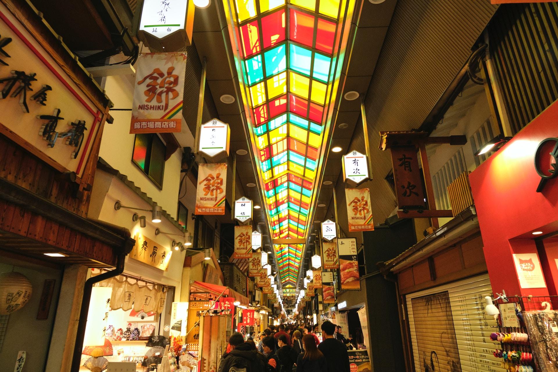 Nishiki Market lit up in the evening with a colourful overhead light panel extending the length of the walkway and the shops lit up with white lights.