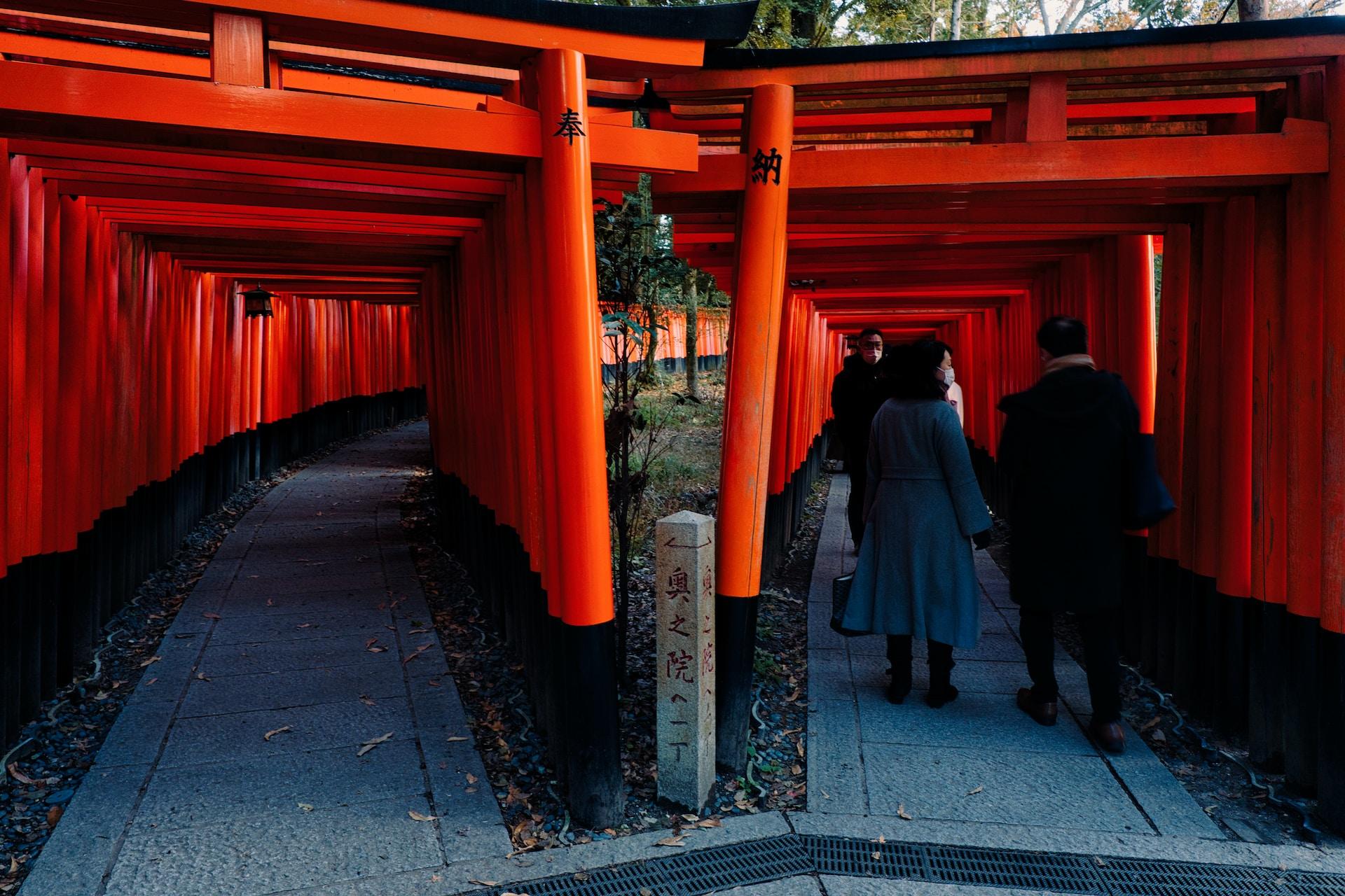A double-pathway access to a Shinto shrine with its framing painted in orange. Three people walk the right-hand path.