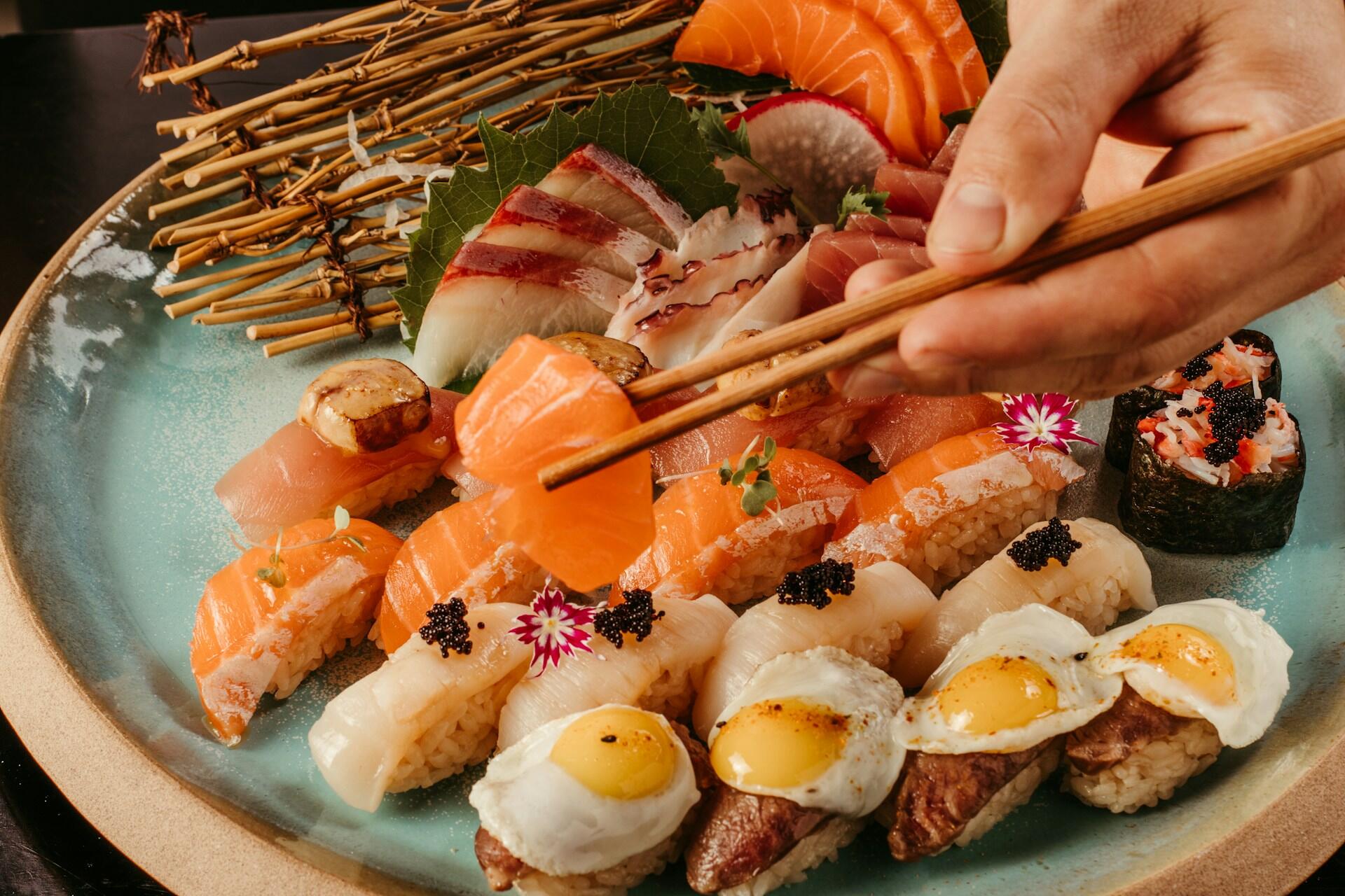 A hand with chopsticks lifts a salmon slice from a vibrant sushi platter.