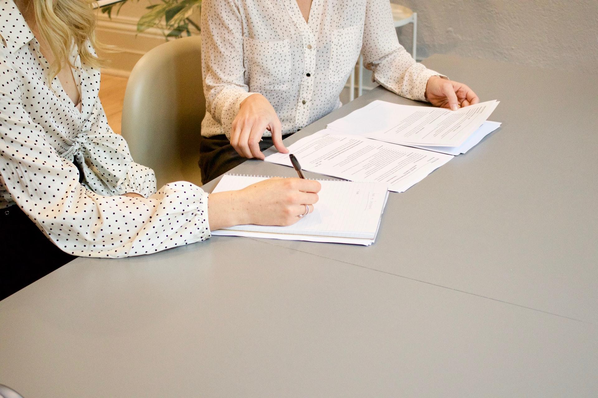A woman in a polka-dotted blouse sits with a woman wearing at tan blouse with pockets, pointing at a sheet of paper while the other woman gathers papers up.